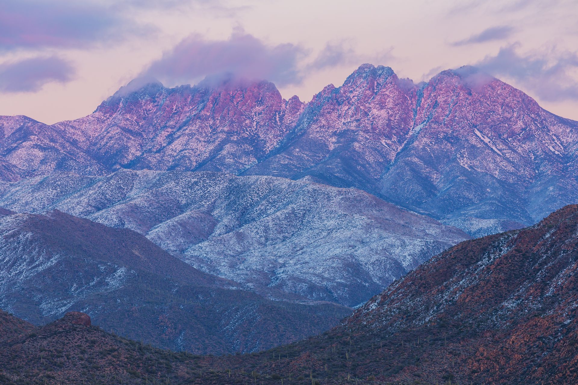 Snowy mountain range at sunset, peaks lit with pink, purple, and lavender hues.