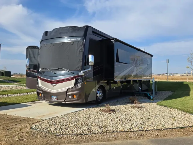 Large brown RV parked on gravel and concrete, with open door and awning, sunny outdoors.