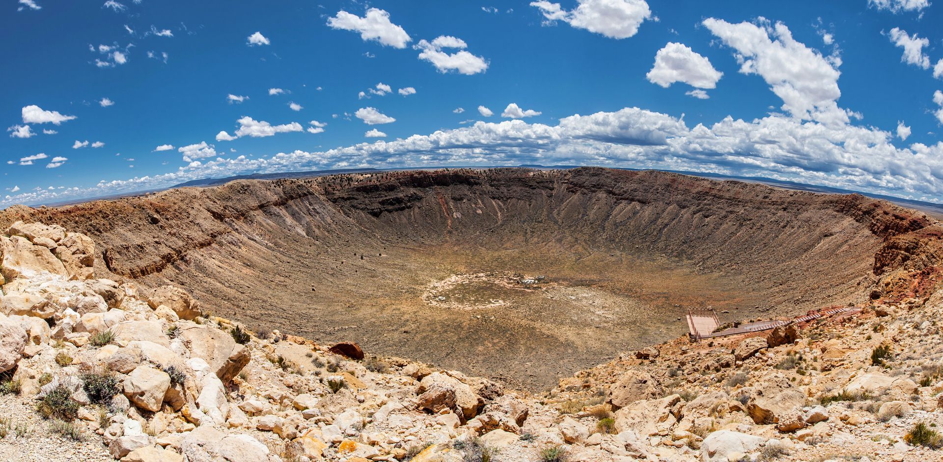 A wide panoramic view of a large, circular crater under a bright blue sky with fluffy white clouds.