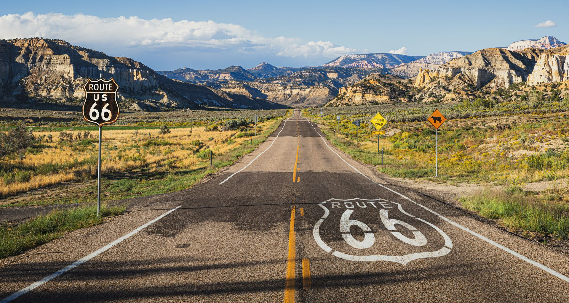 Route 66 highway stretches through a desert landscape, marked by road signs and white painted road symbols.