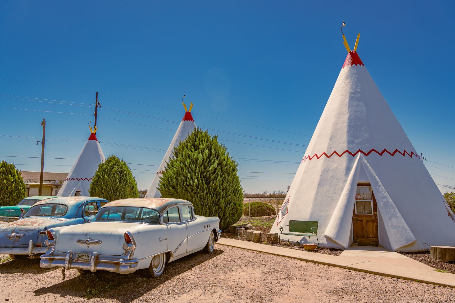 Tepee-shaped motel units with vintage cars parked outside, set against a blue sky.