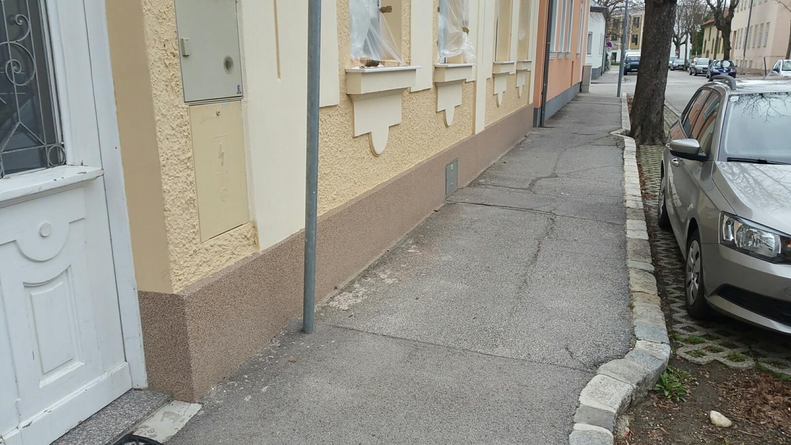 Ein Gehweg verläuft entlang eines cremefarbenen Gebäudes mit strukturiertem Sockel, neben dem ein silbernes Auto auf der Straße geparkt ist.