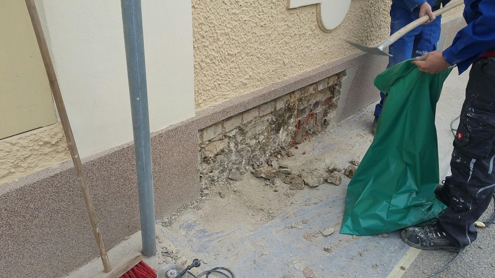 Ein Bauarbeiter in Arbeitshose hält einen grünen Bauschuttsack in der Hand, während er einen beschädigten Abschnitt eines Hausfundaments reinigt.