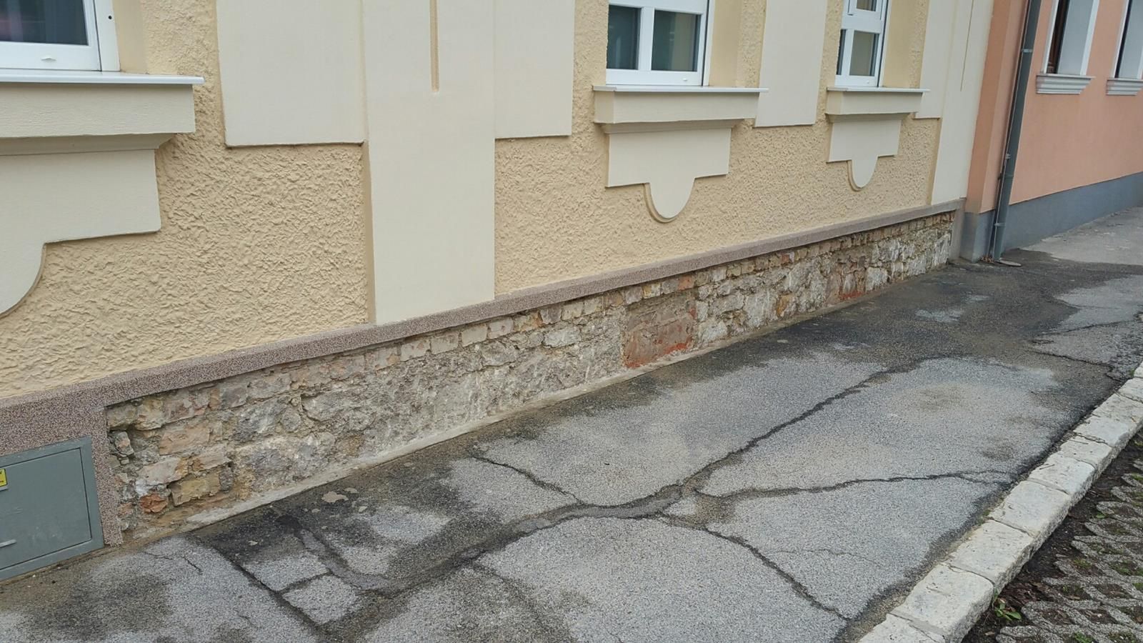 Eine beigefarbene Gebäudefassade mit beschädigtem Steinfundament und rissigem Gehwegpflaster.