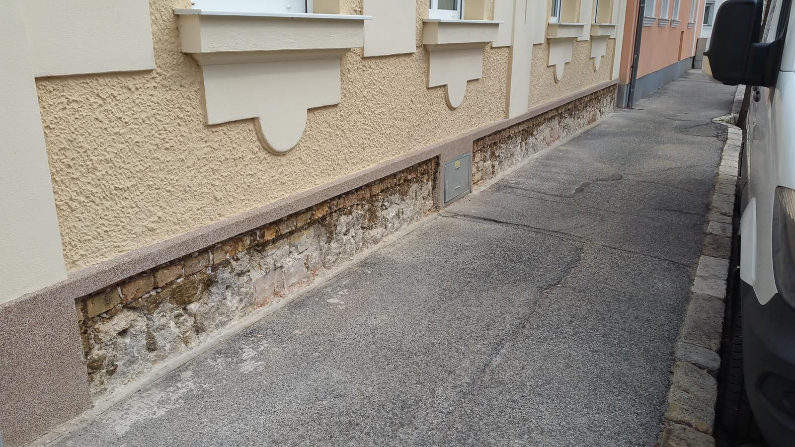 Eine Gebäudemauer mit beschädigtem, freiliegendem Ziegelsockel entlang eines schmalen gepflasterten Gehwegs.