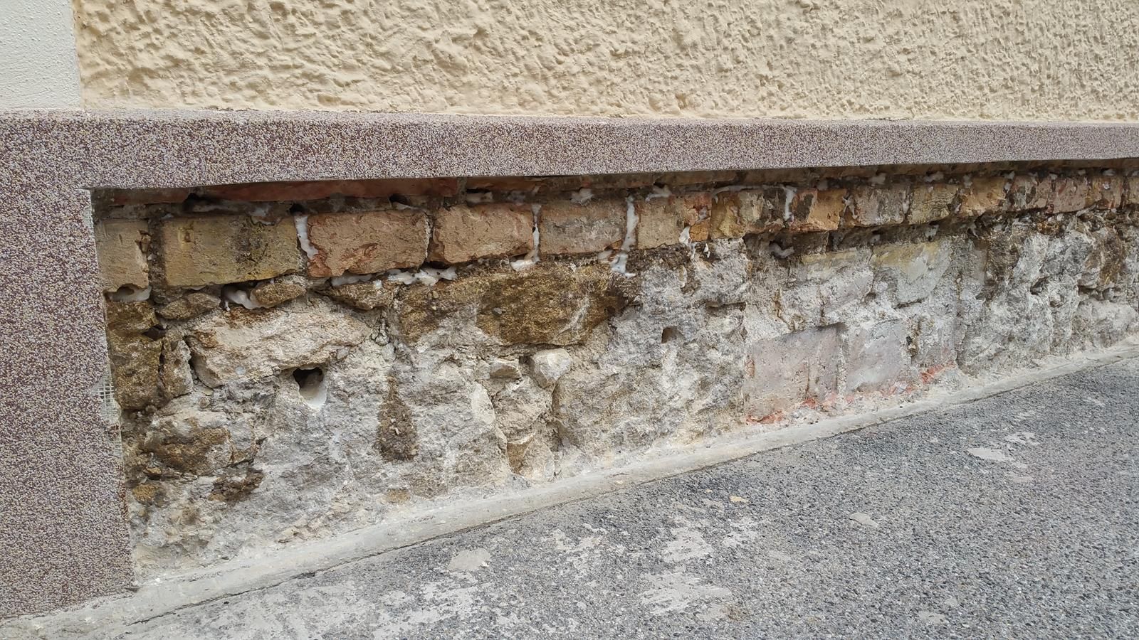 Ein äußeres Gebäudefundament mit freiliegendem, bröckelndem Ziegel- und Steinmauerwerk unter einer glatten Steinverkleidung und -mauer.