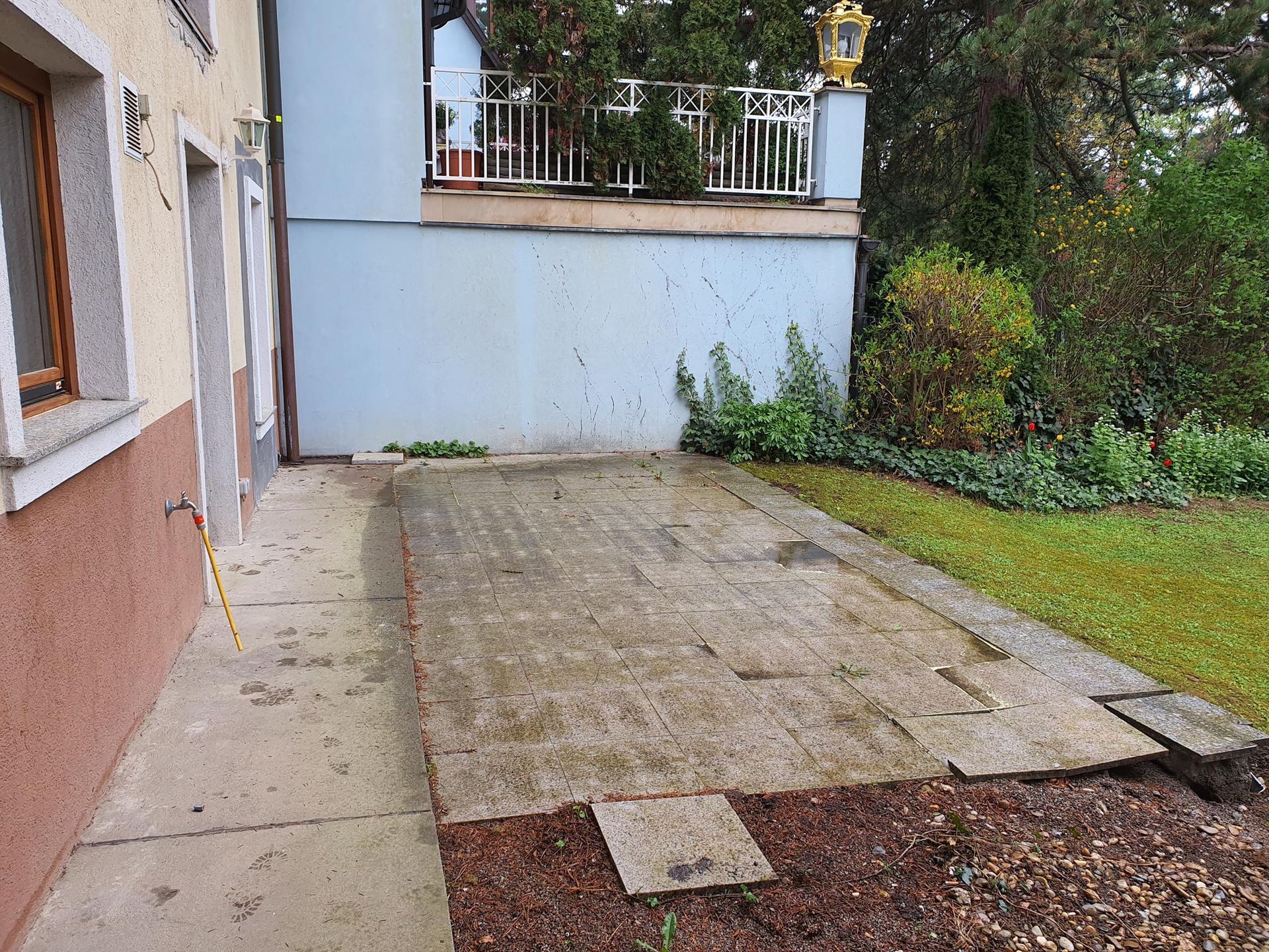 Eine helle, mit Steinplatten belegte Terrasse grenzt an eine Hauswand und einen erhöhten Balkon mit weißem Geländer.