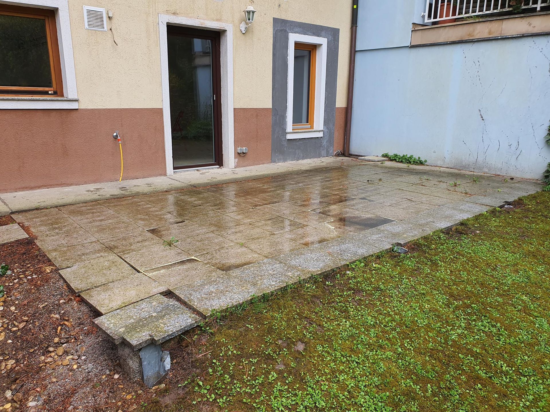 Neben einem hellfarbigen Haus mit dunkel gerahmten Türen und Fenstern befindet sich eine rechteckige Steinterrasse, angrenzend an eine Rasenfläche.