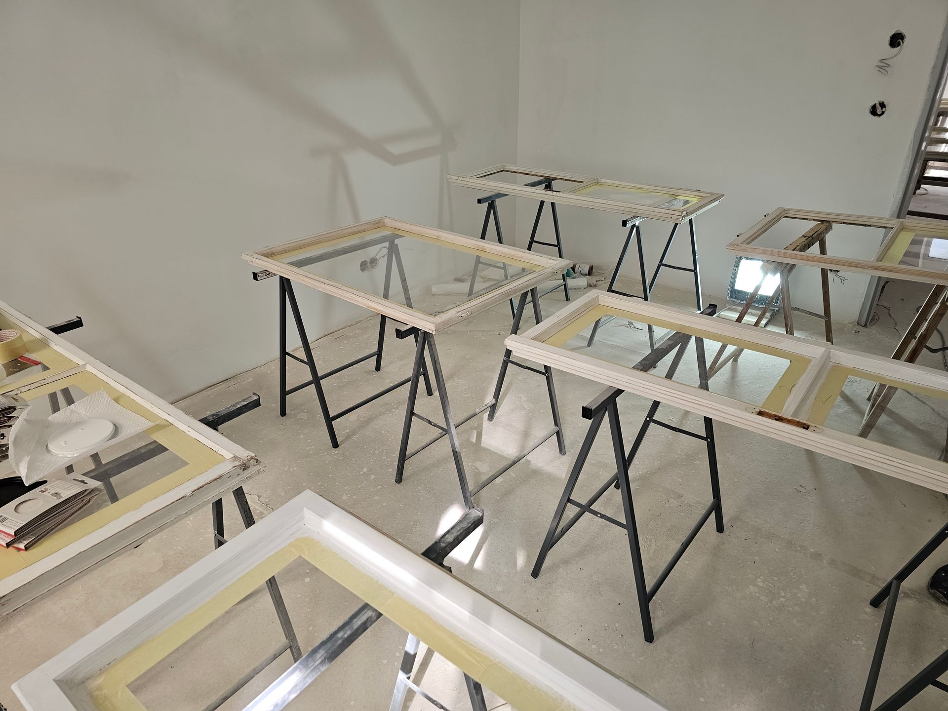 Mehrere weiße Fensterrahmen ruhen auf schwarzen Sägeböcken in einem hell erleuchteten, leeren Raum.