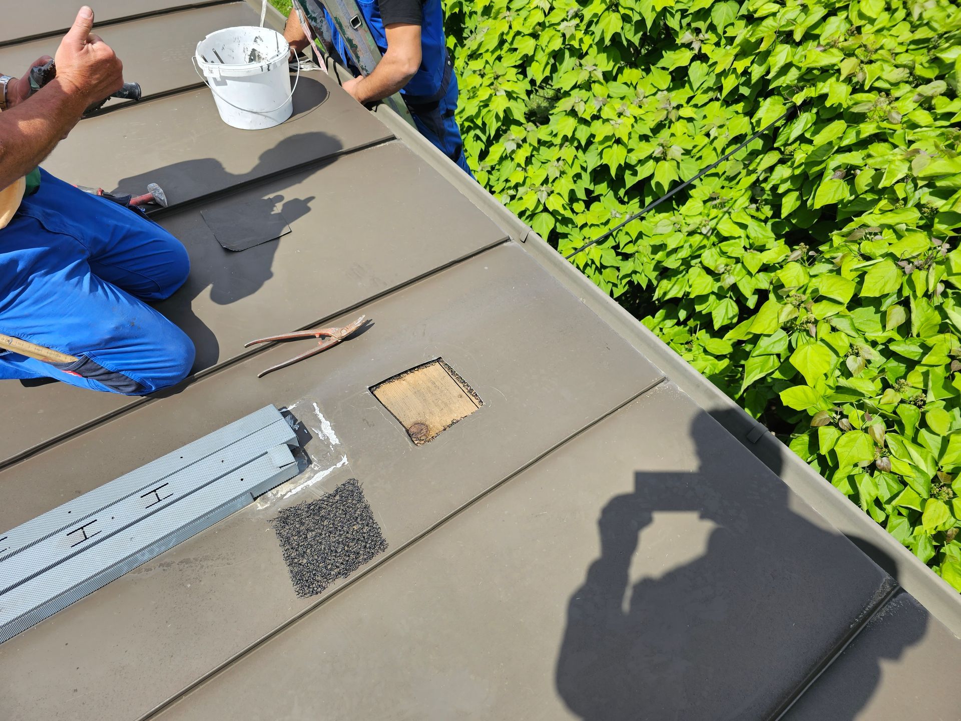 Zwei Arbeiter in blauen Arbeitskleidungen reparieren ein Metalldach; Werkzeug und ein weißer Eimer stehen daneben, neben einer üppigen grünen Hecke.