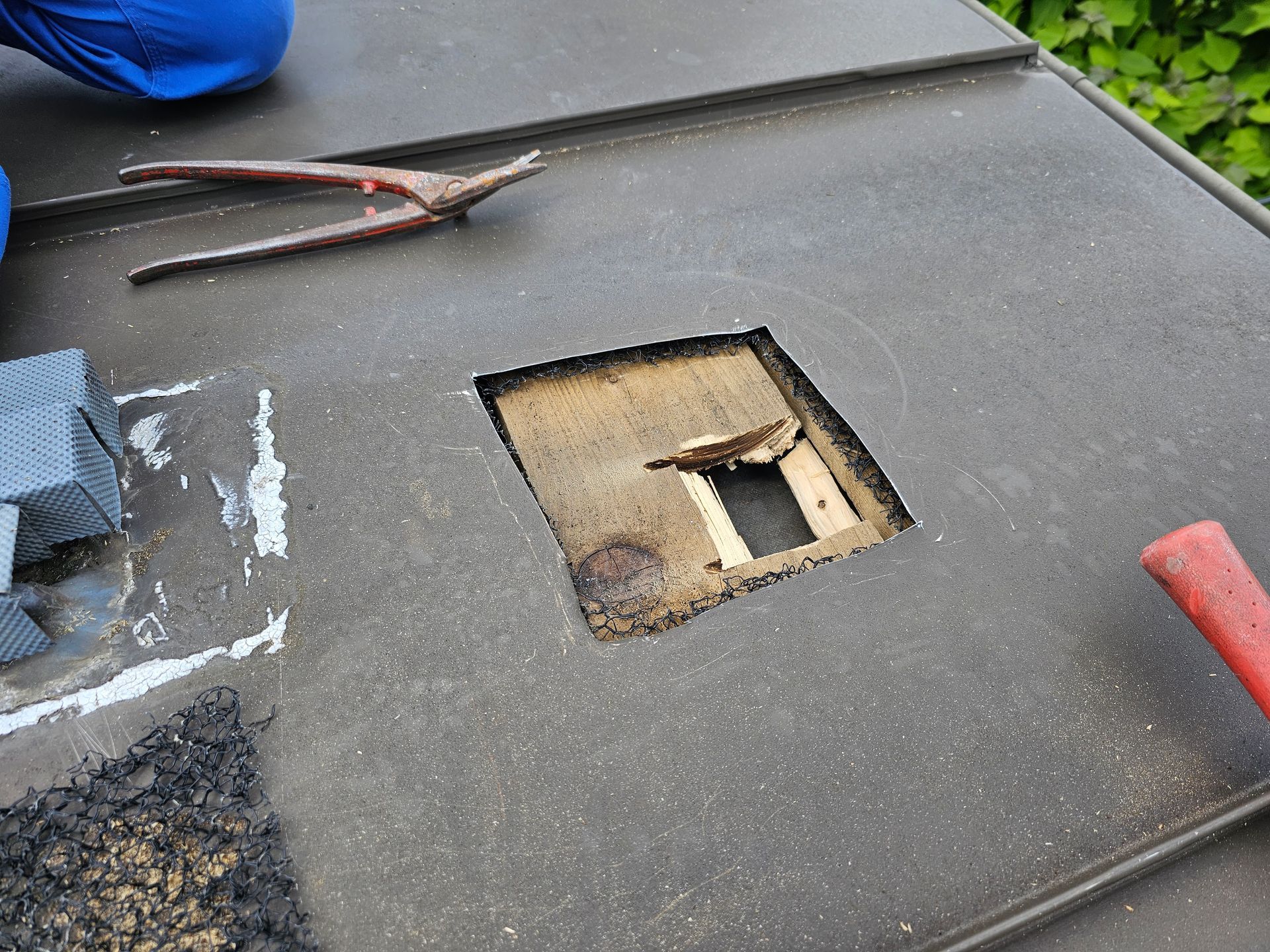 In die dunklen, strukturierten Dachschindeln ist ein quadratisches Loch geschnitten, das die hölzerne Dachkonstruktion freilegt; Werkzeuge für Reparaturen liegen daneben.