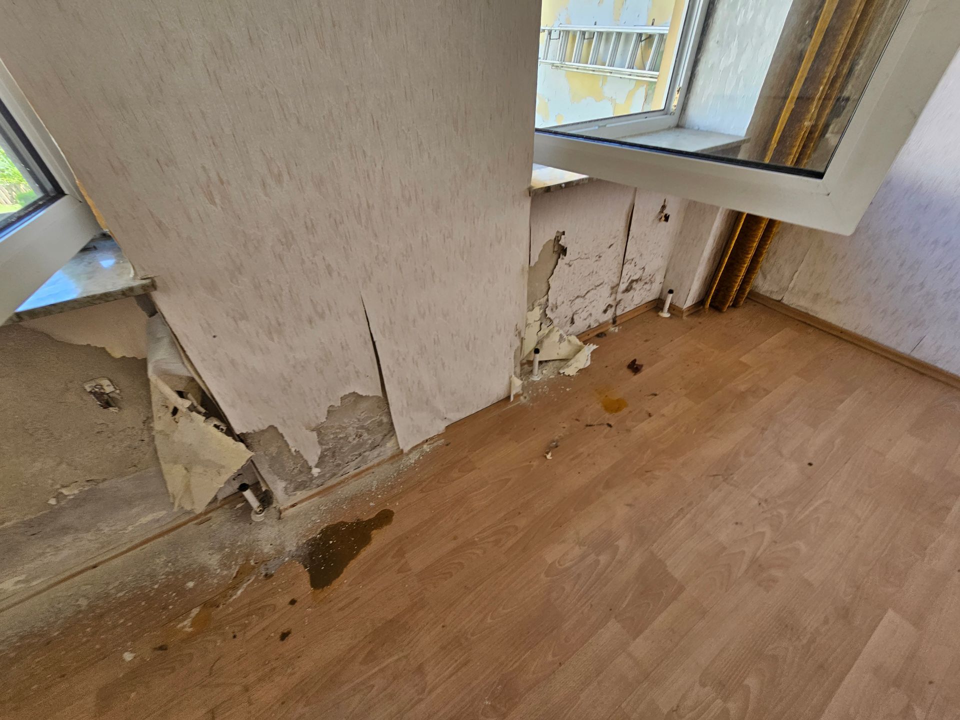 Wasserschäden und abblätternde Tapete um einen Fensterrahmen herum sowie nasse, fleckige Dielen in einem Zimmer.