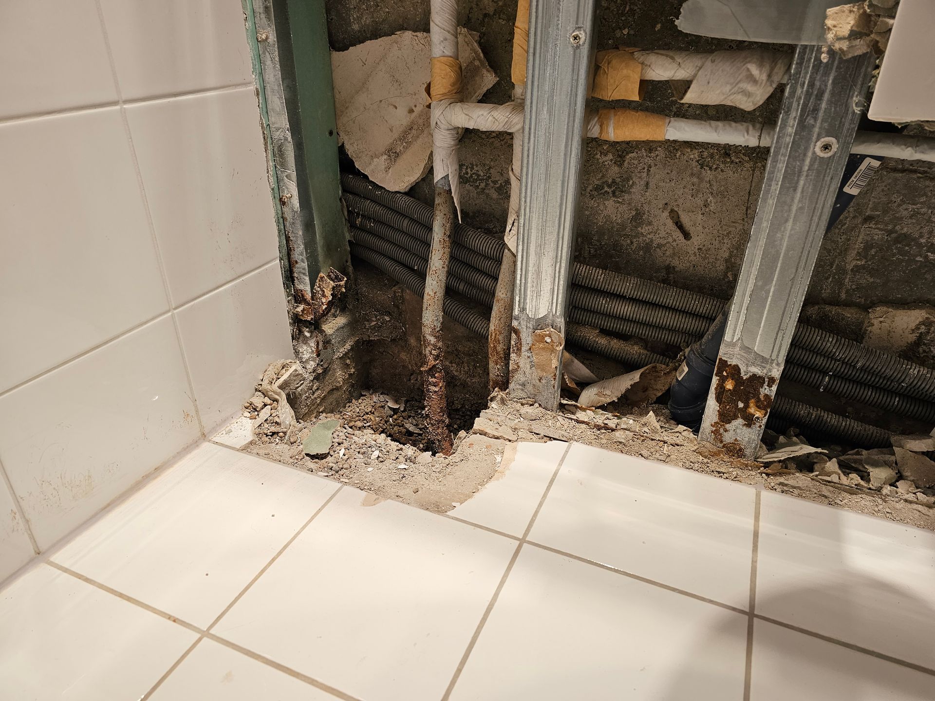 Ein Wandabschnitt wurde geöffnet und gab den Blick frei auf freiliegende Metallrahmen und Wasserleitungen mit sichtbarer Korrosion hinter weißen Bodenfliesen.
