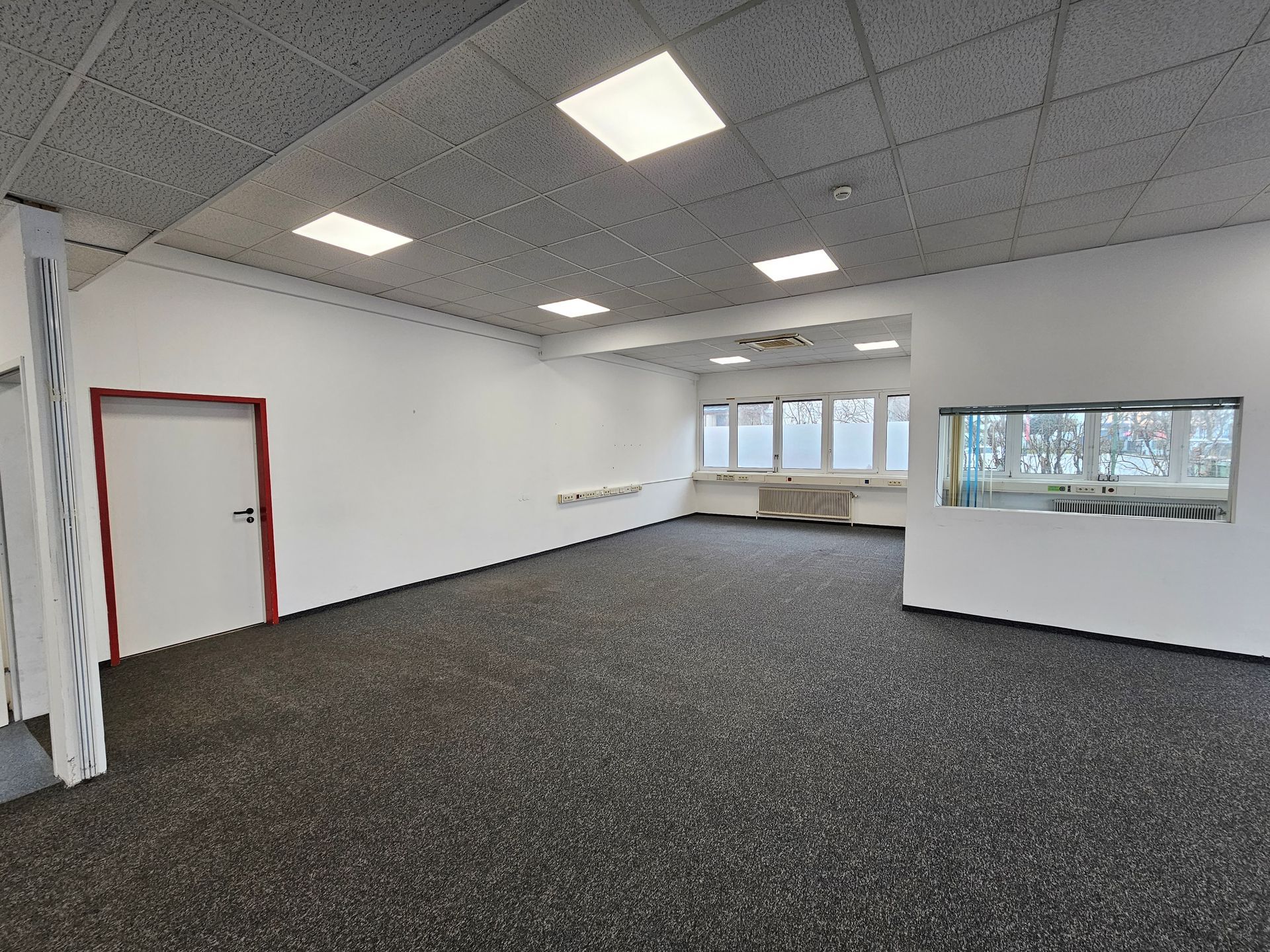 Ein leerer, geräumiger Büroraum mit weißen Wänden, dunkelgrauem Teppichboden, Deckeneinbaustrahlern und zwei Fensterfronten.