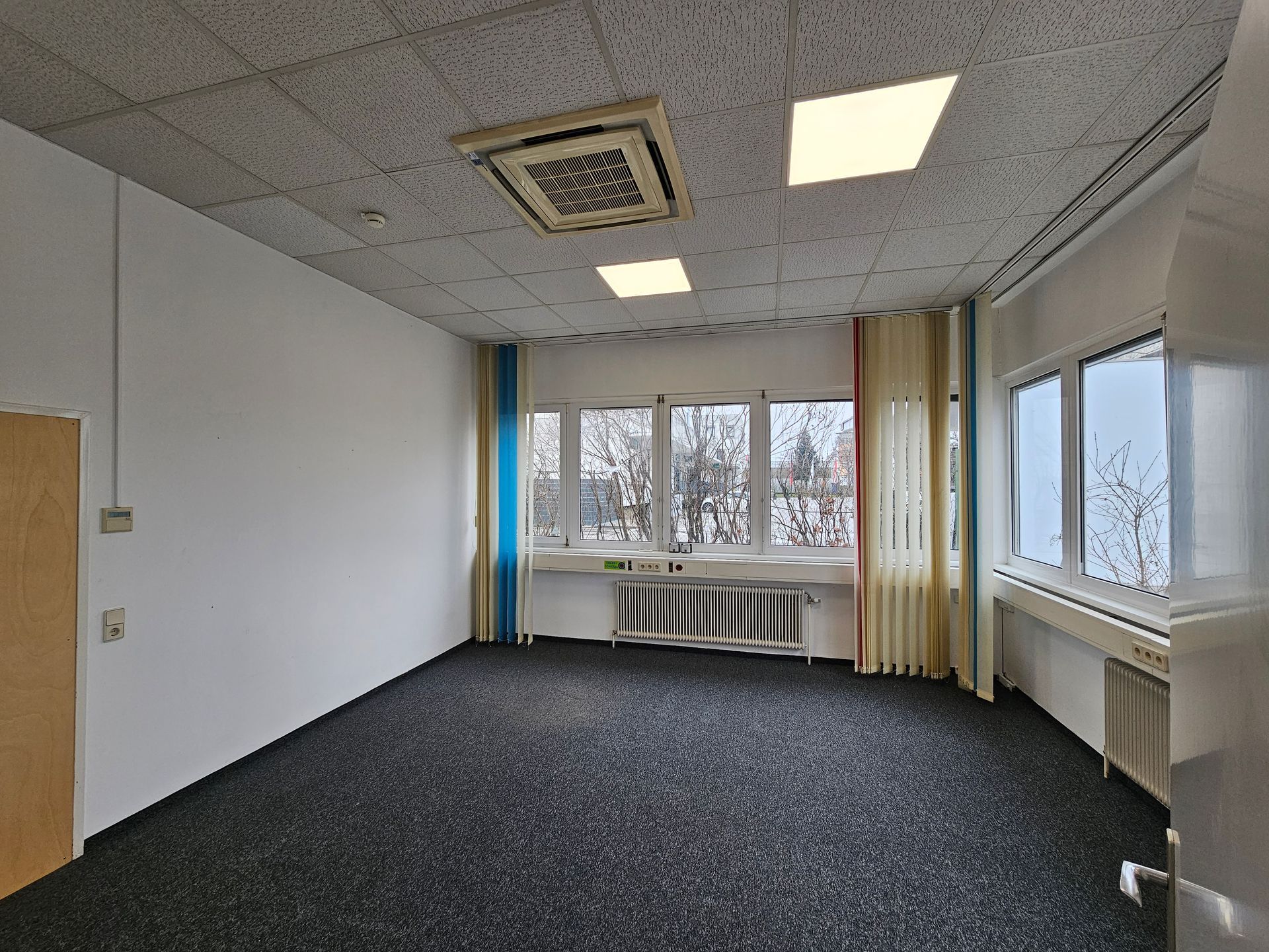 Ein leeres Büro mit weißen Wänden, dunkelgrauem Teppichboden, zwei Deckenleuchten, Fenstern mit Vorhängen und einem Heizkörper.