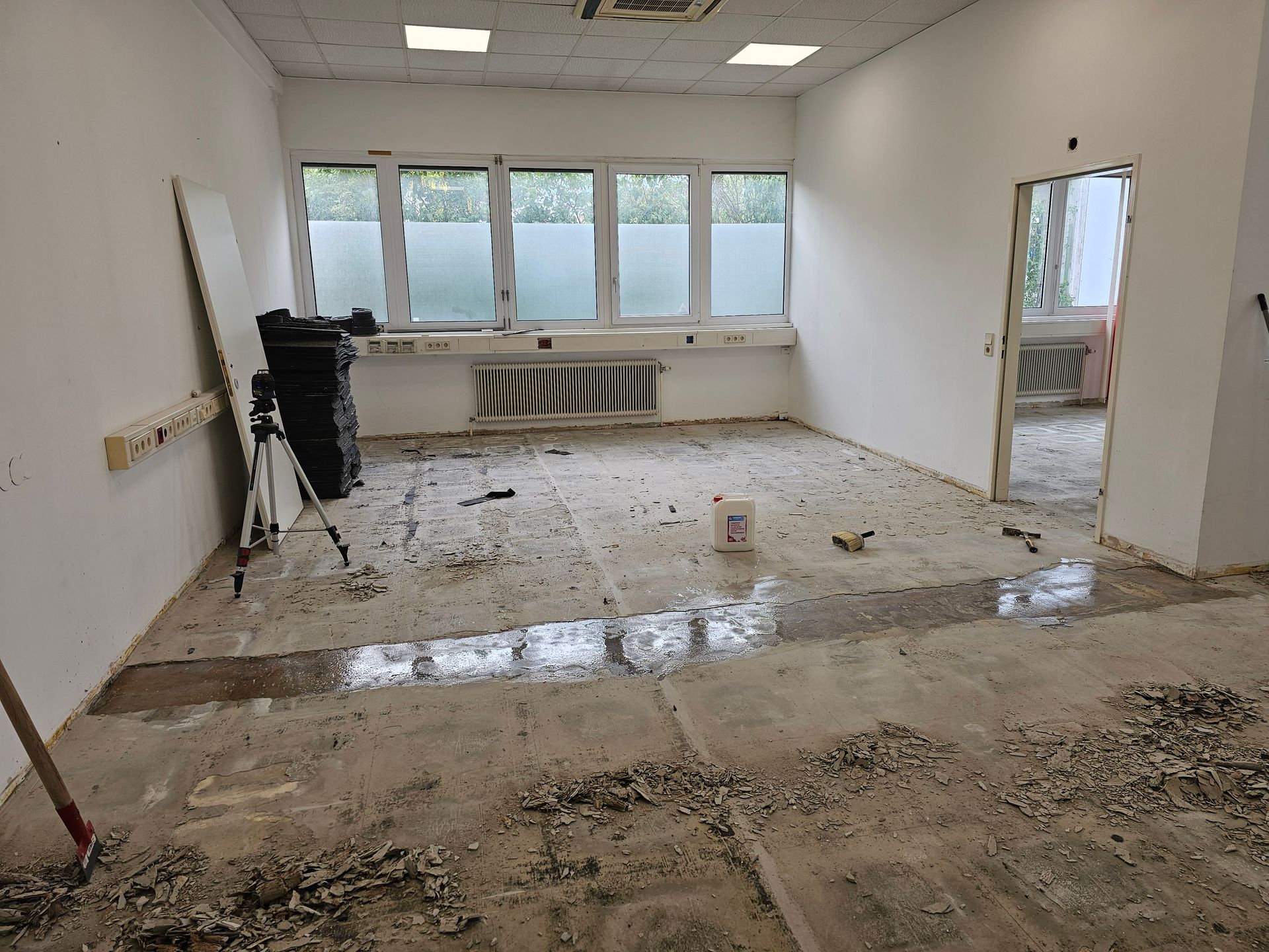 Ein leerer, weiß getünchter Raum, der gerade renoviert wird, mit Bauschutt auf dem Boden und einem Stativ in der Nähe eines Fensters.