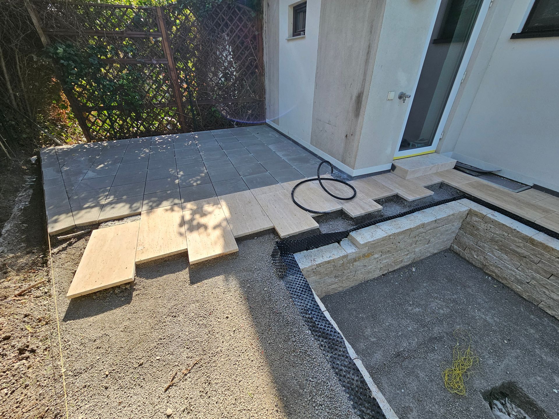 Ein im Bau befindlicher Terrassenbereich im Freien mit grauen Pflastersteinen, hellen Steinfliesen und einer mit Kies gefüllten Grube.