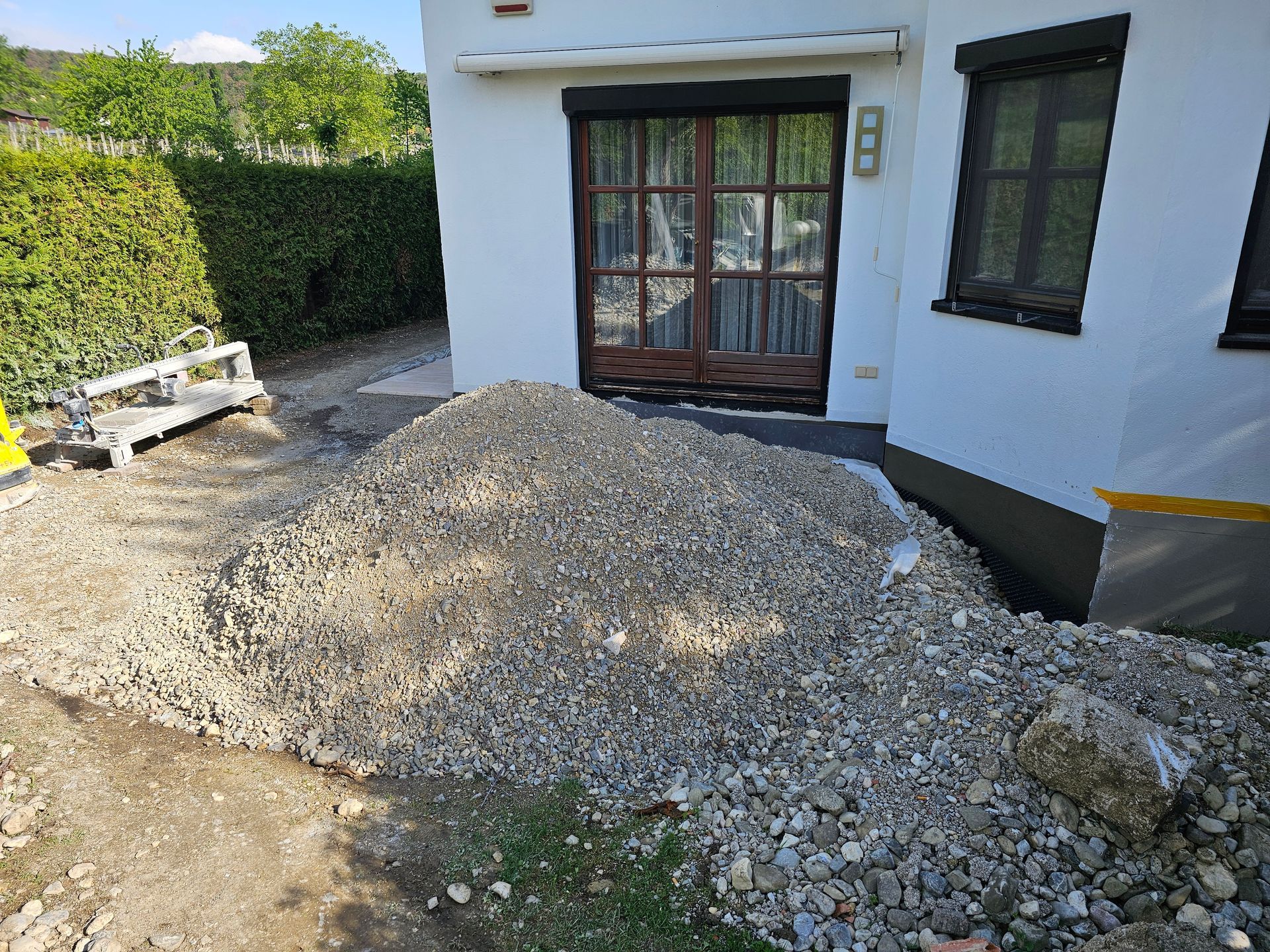 Vor einem weißen Haus mit Terrassentür und einer Hecke an der Seite liegt ein Haufen Kies auf dem Boden.