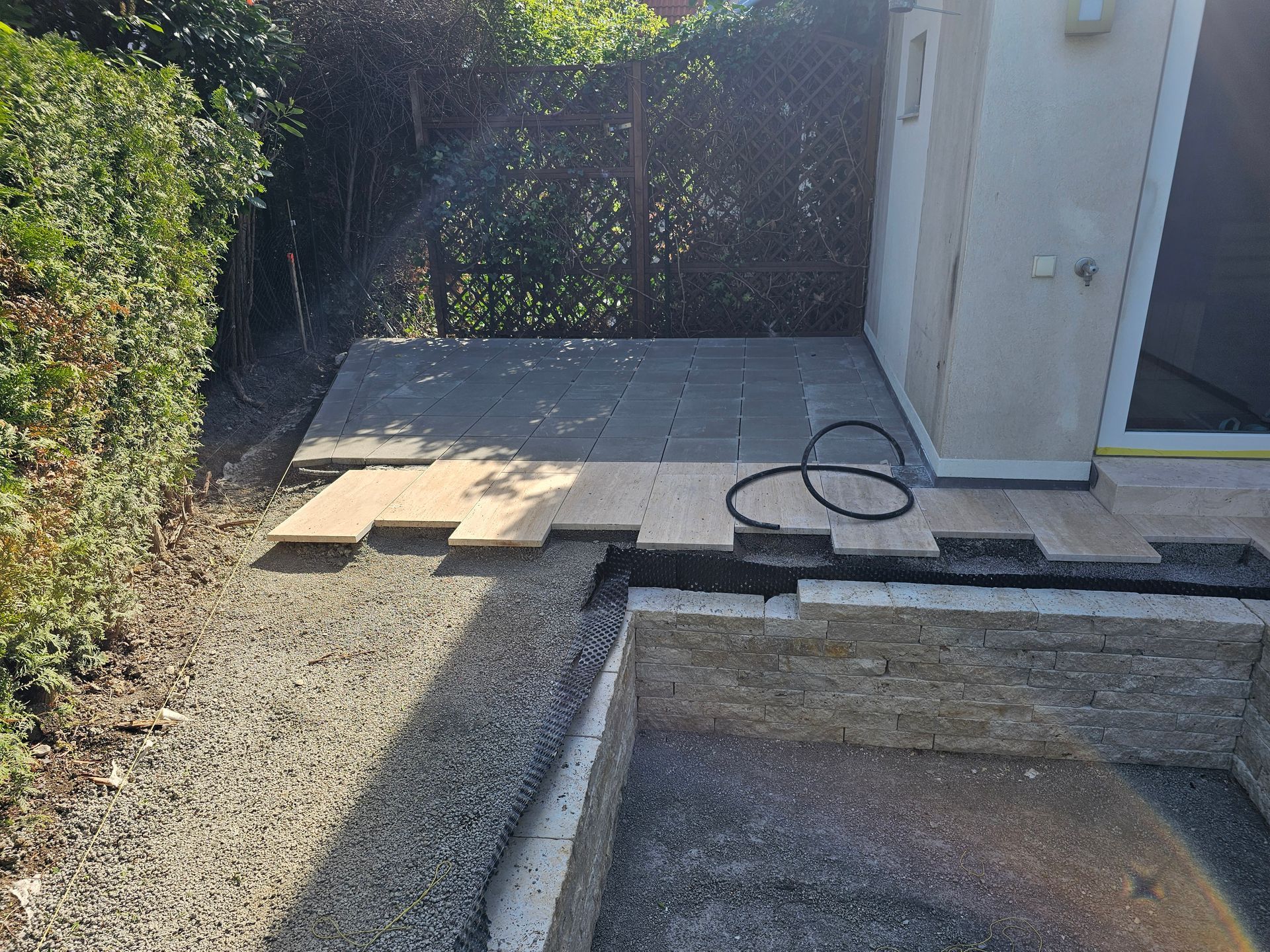 Ein im Bau befindlicher Hinterhof-Terrassenbereich mit grauen Steinfliesen, Holzplanken, einem Kiesweg und einer Steinstützmauer.