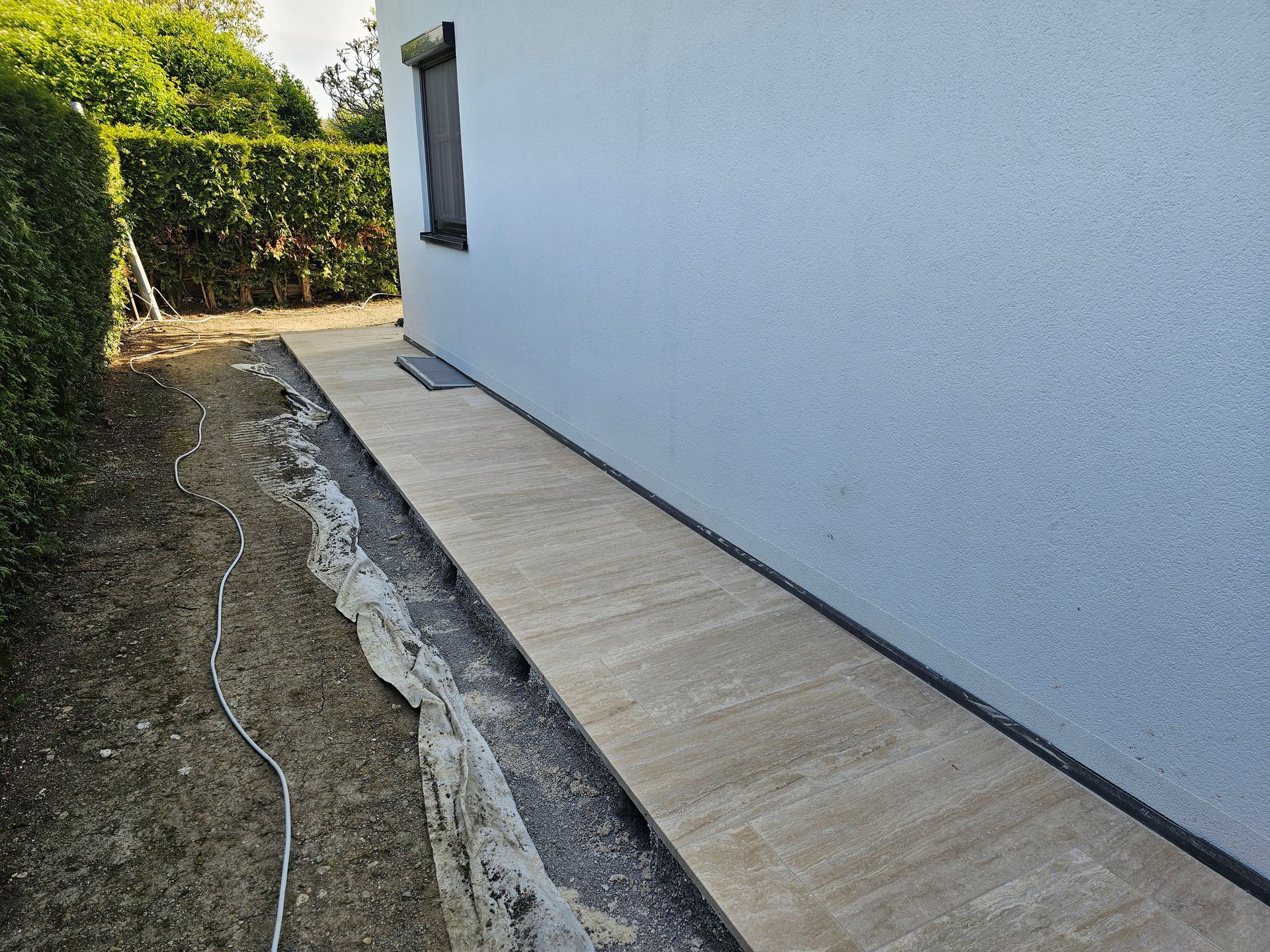 Ein heller Steinweg verläuft an der Seite eines modernen weißen Hauses neben einer dunklen Hecke und einem unbestellten Boden.