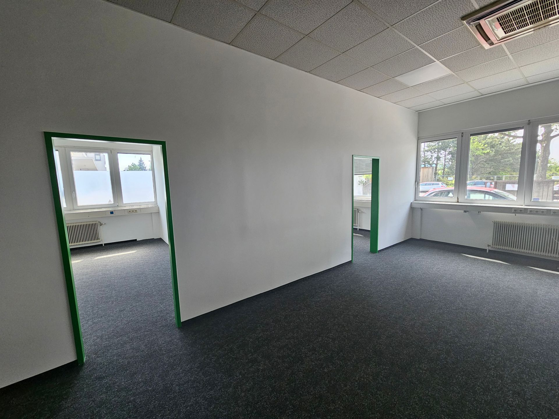 Innenansicht eines hellen Büroraums mit dunkelgrauem Teppichboden, weißen Wänden, grün gerahmten Türrahmen und großen Fenstern.