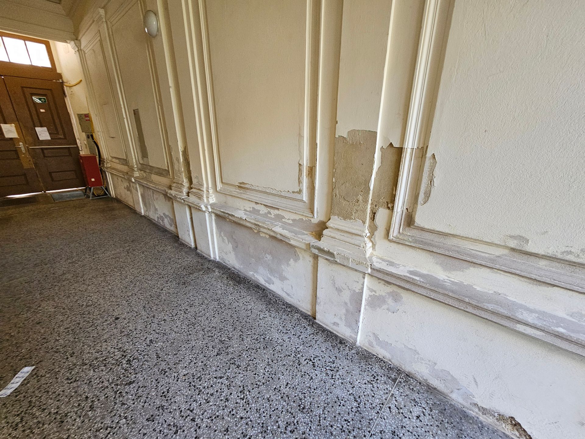 Ein Flur mit gesprenkeltem Steinboden und einer cremefarbenen Wand mit Zierleisten und beschädigter, abblätternder Farbe.