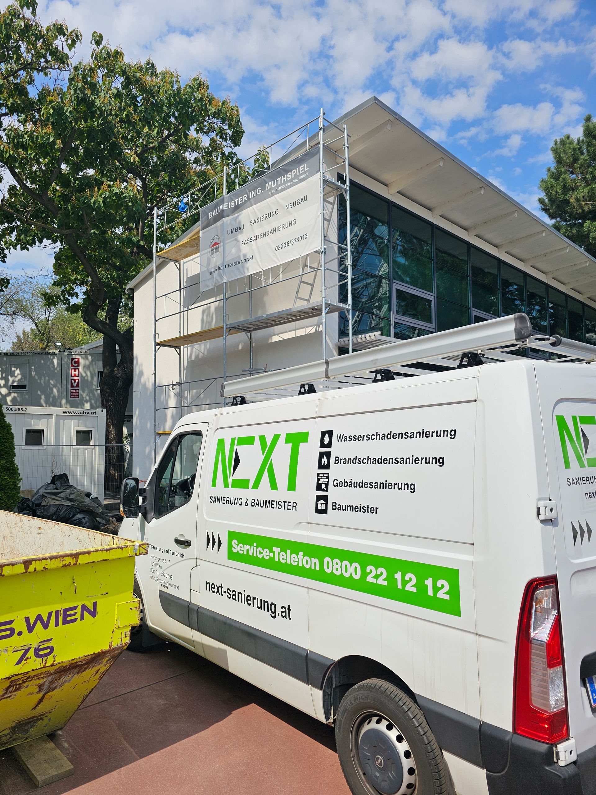 Ein weißer Lieferwagen mit grüner „NEXT“-Aufschrift parkt an einem sonnigen Tag vor einem eingerüsteten Gebäude.