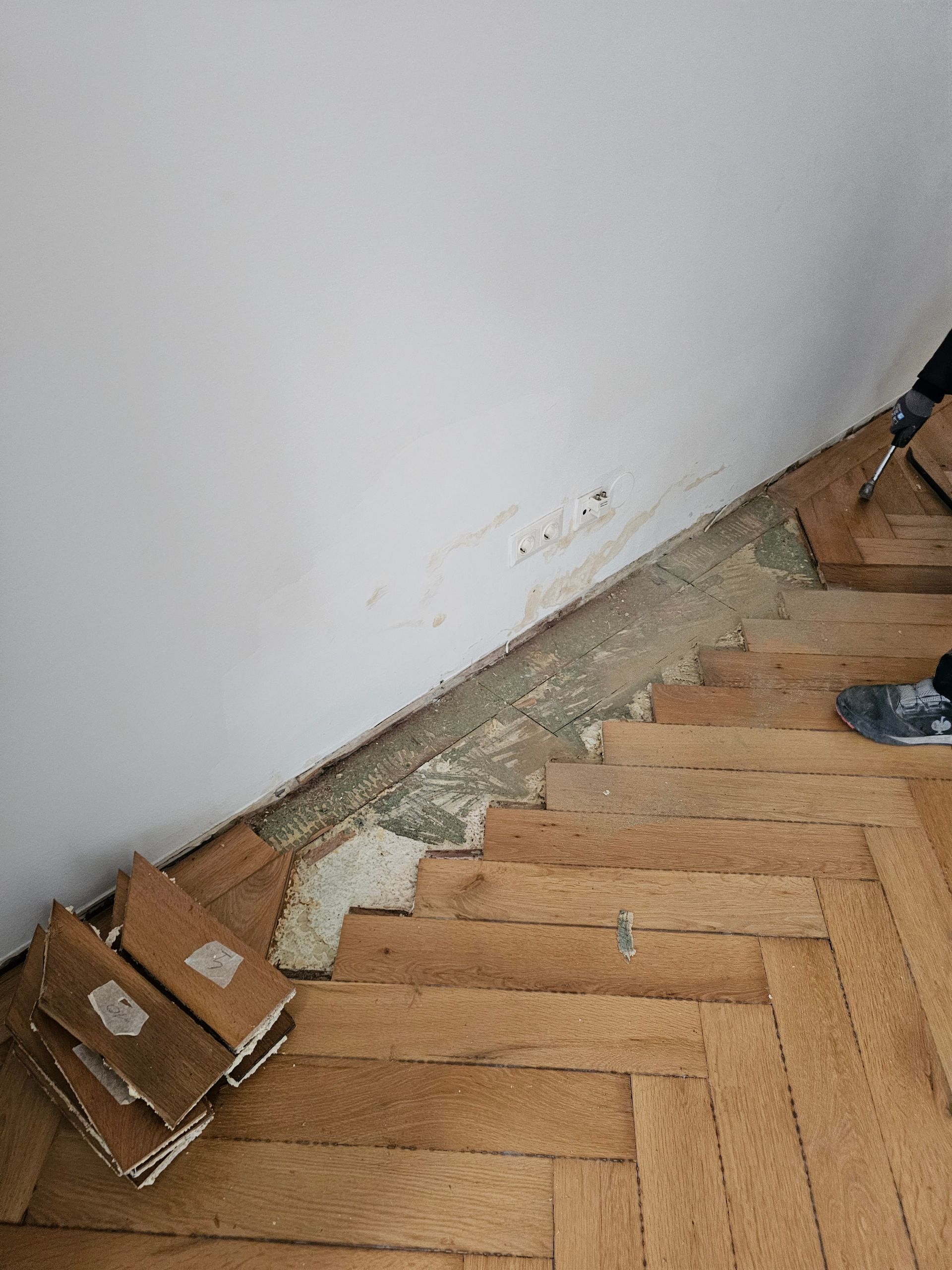 Ein menschlicher Fuß und ein Werkzeug entfernen einen Holzparkettboden und lassen dabei eine freiliegende Betonkante an einer weißen Wand zurück.