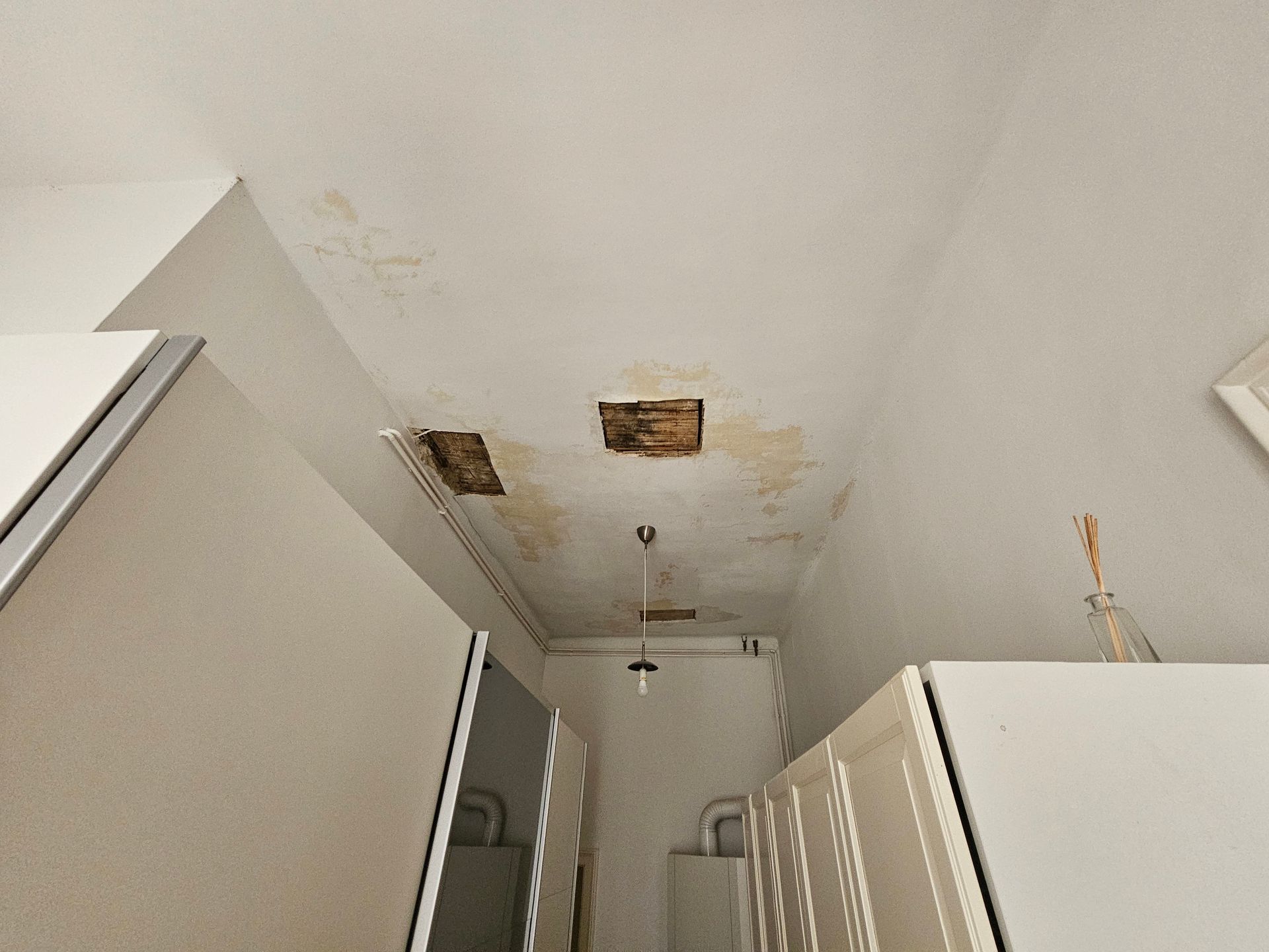 Eine beschädigte Decke mit großen gelben Wasserflecken, abblätternder Farbe und zwei Löchern, die sich über den Küchenschränken befindet.