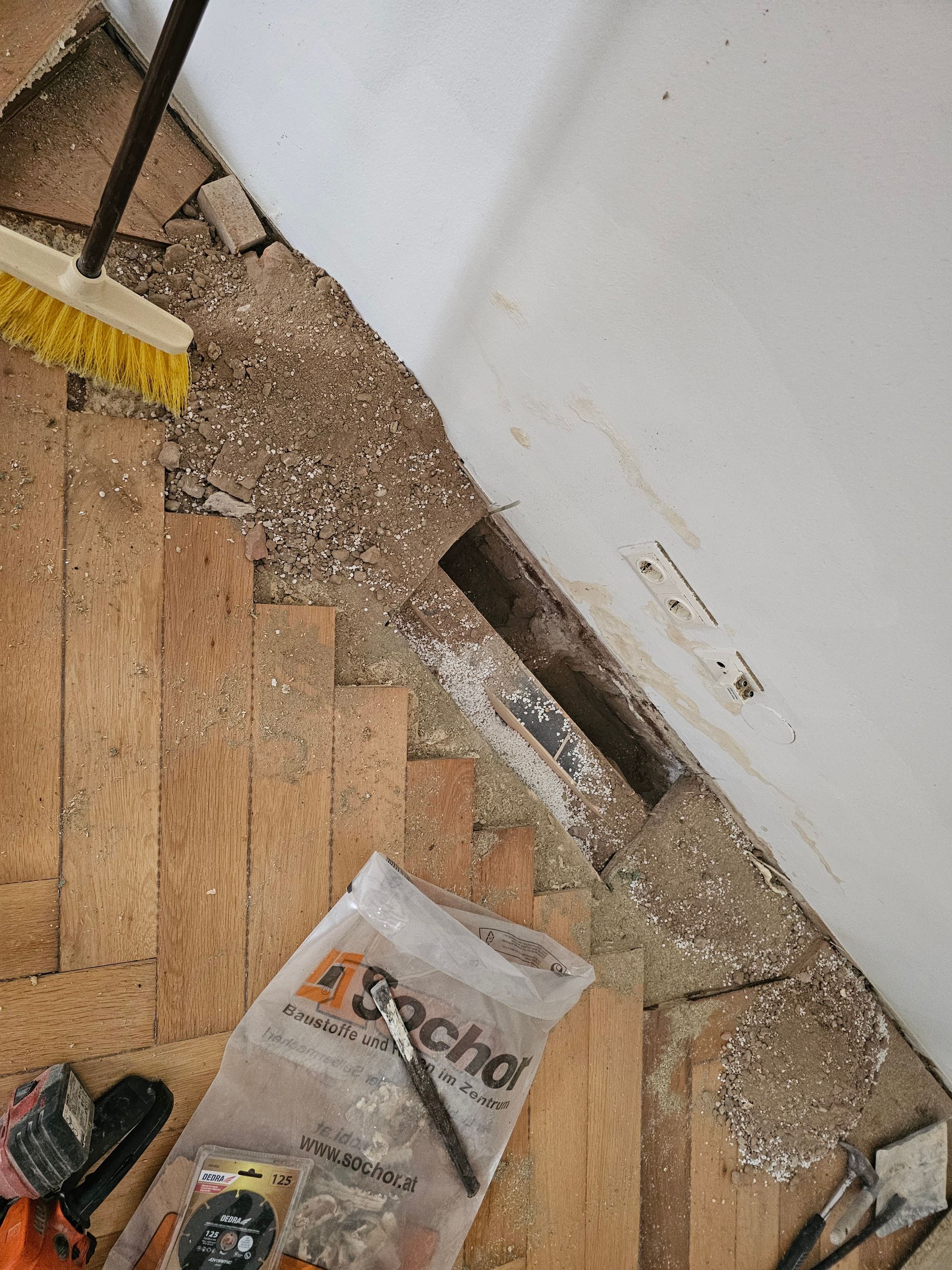 Eine Baustelle mit staubigem Boden und einem rechteckigen Lüftungsausschnitt in der Nähe einer Wand, daneben Werkzeuge und eine Materialtasche.