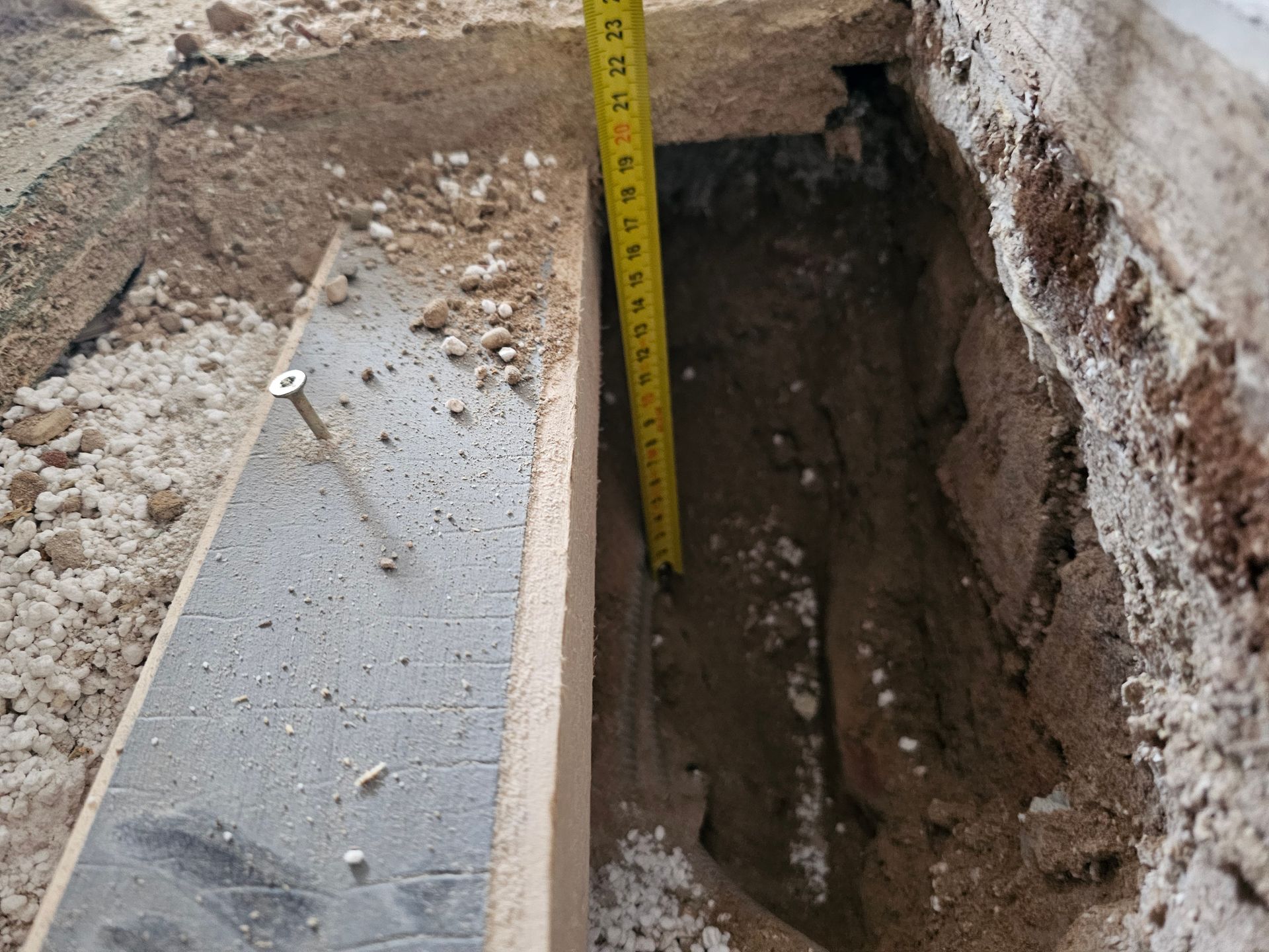 In einem schmalen, frisch ausgehobenen Erdgraben neben einer Bauschalung und einem Steinfundament steht ein Maßband senkrecht.