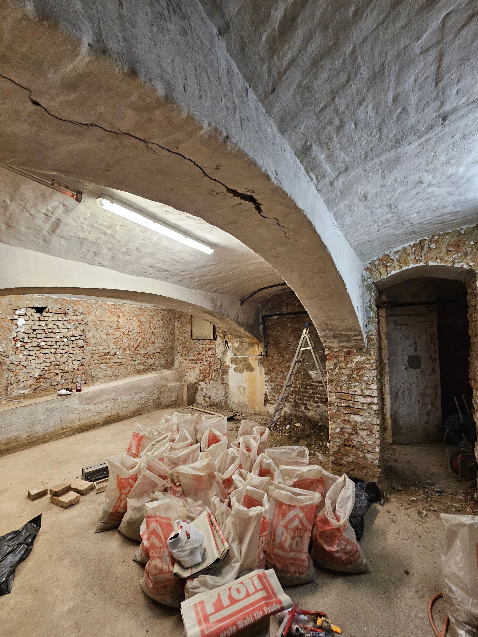 Ein Kellerraum mit gewölbten Steindecken, freiliegenden Ziegelwänden und Säcken voller Baumaterialien auf dem Boden.