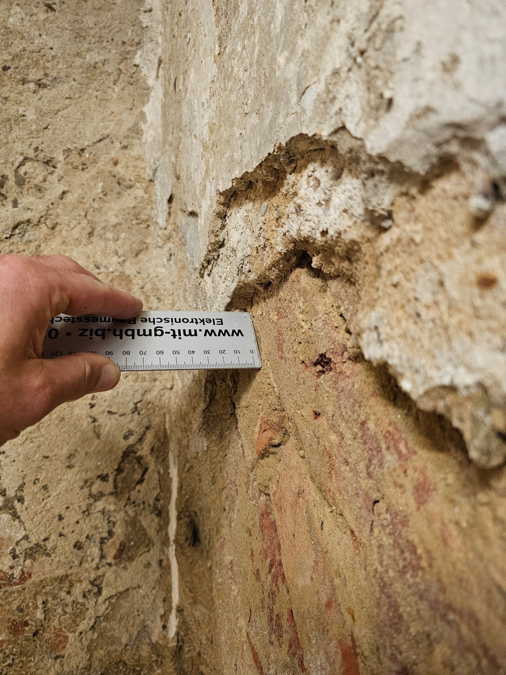Eine Hand hält ein Lineal an eine beschädigte, strukturierte Mauer, an der Schichten aus Putz und Ziegeln sichtbar sind.