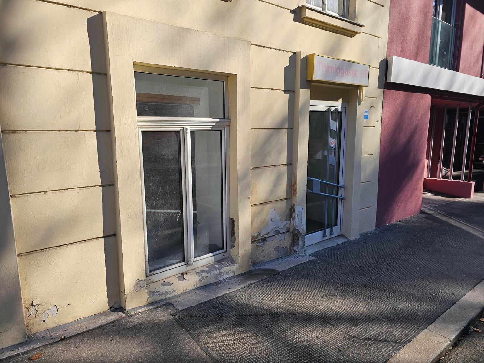 Eine cremefarbene Gebäudefassade mit Fenster und Glastür an einem Gehweg. Die untere Wand weist sichtbare Schäden auf.