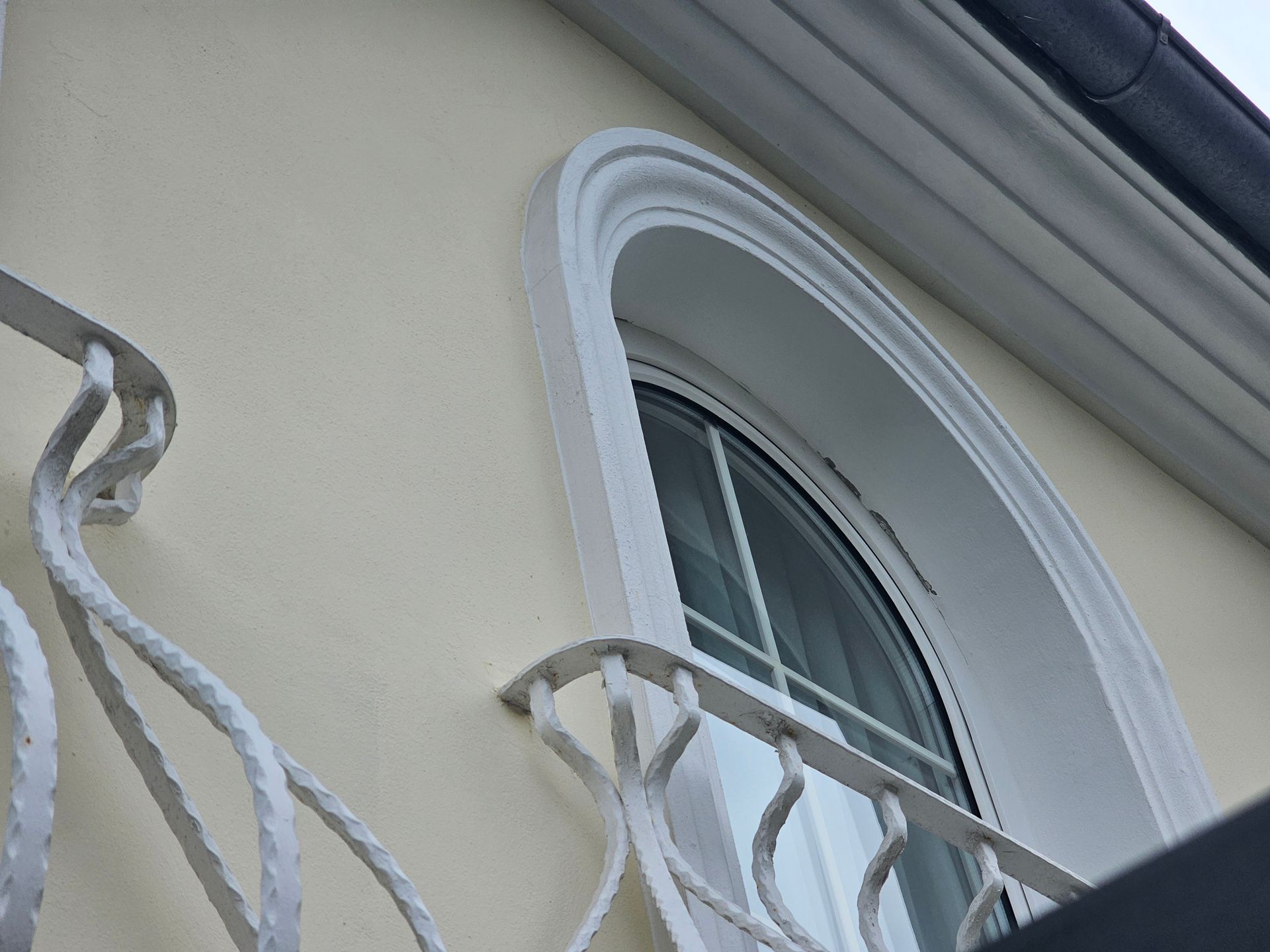 Ein Bogenfenster mit weißem Rahmen an einem hellen Gebäude, gesehen hinter einem kunstvollen schmiedeeisernen Balkongeländer.