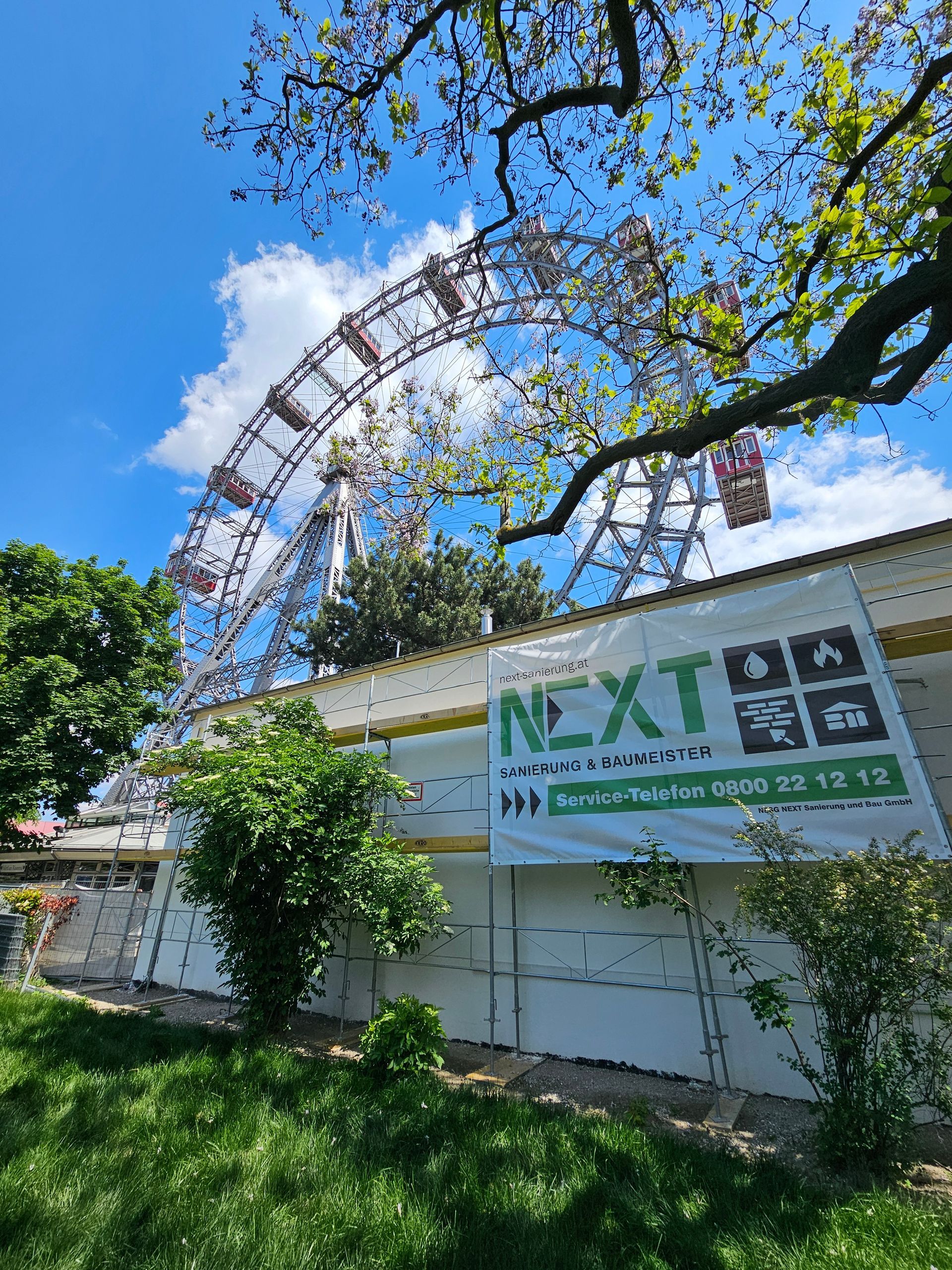 Im Wiener Prater ragt das Riesenrad über einen Bauzaun mit einem grün-weißen „NEXT“-Banner empor.