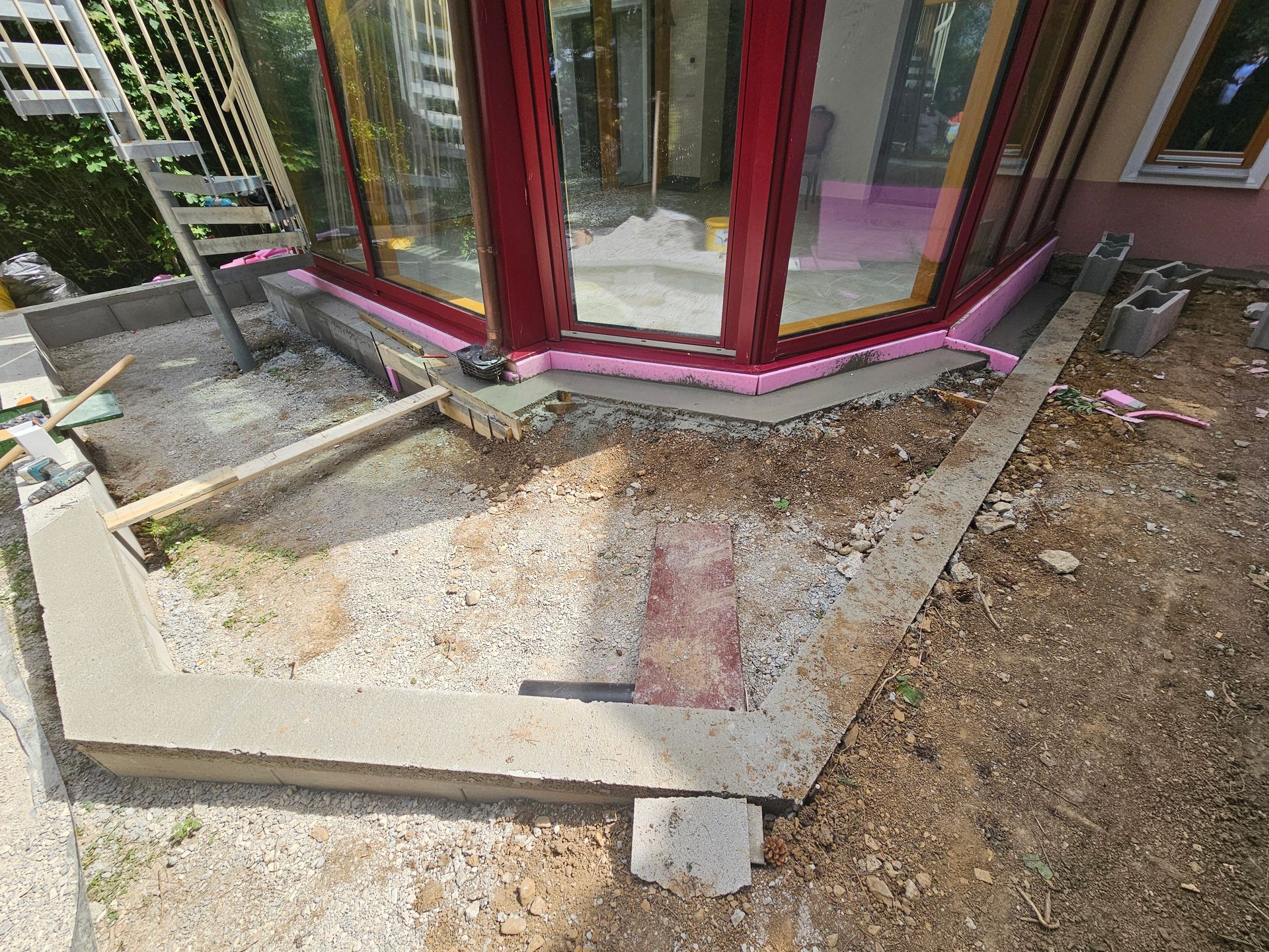 Eine Baustelle mit einer Betonfundamentmauer, die eine mit Kies bedeckte Terrasse vor einem Haus mit roter Umrandung umschließt.