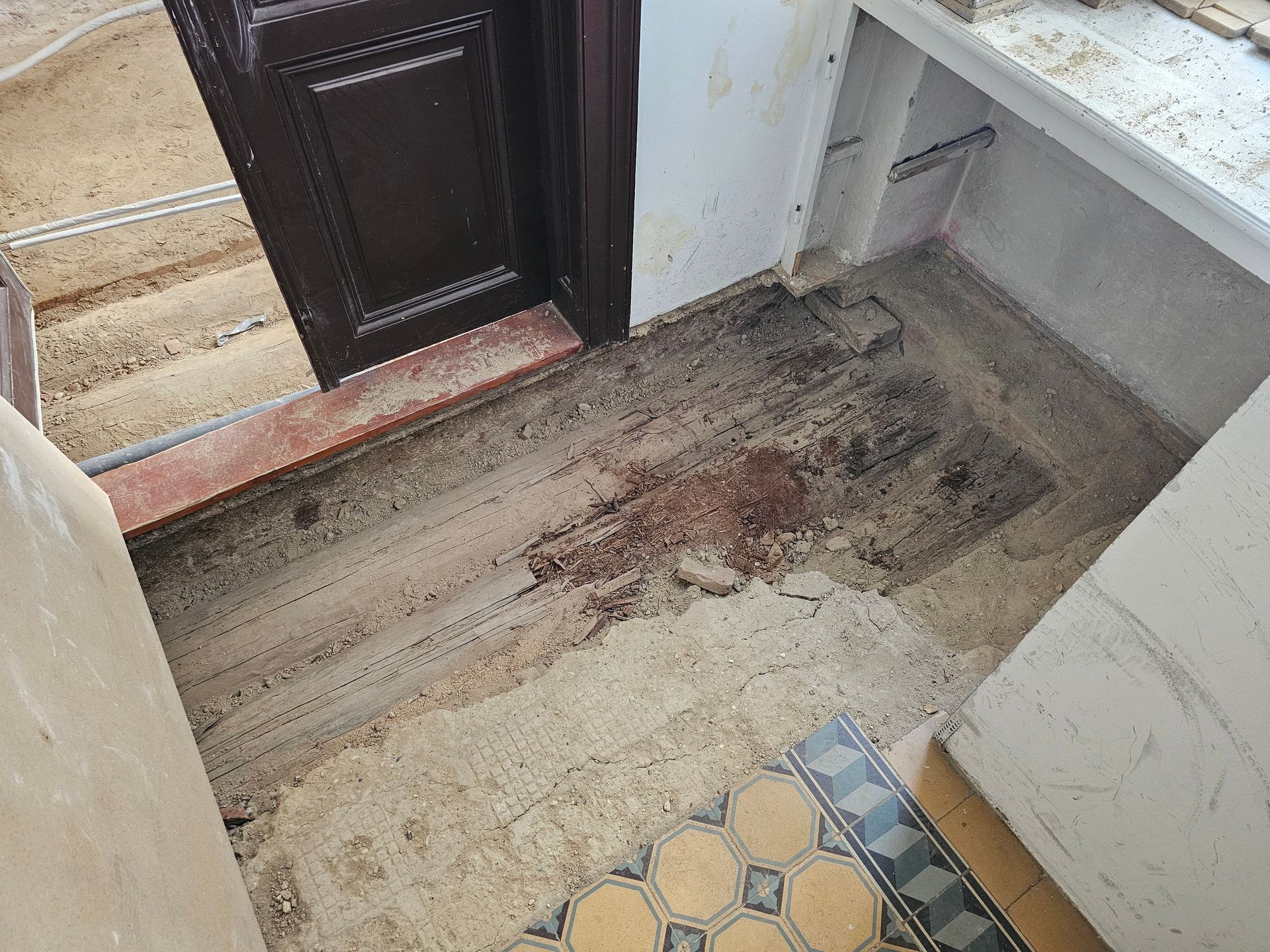 Ein beschädigter Fußboden mit freiliegendem Beton, dunklen Trümmern und teilweise abgenutzten Fliesen in der Nähe einer Holztür und einer weißen Theke.