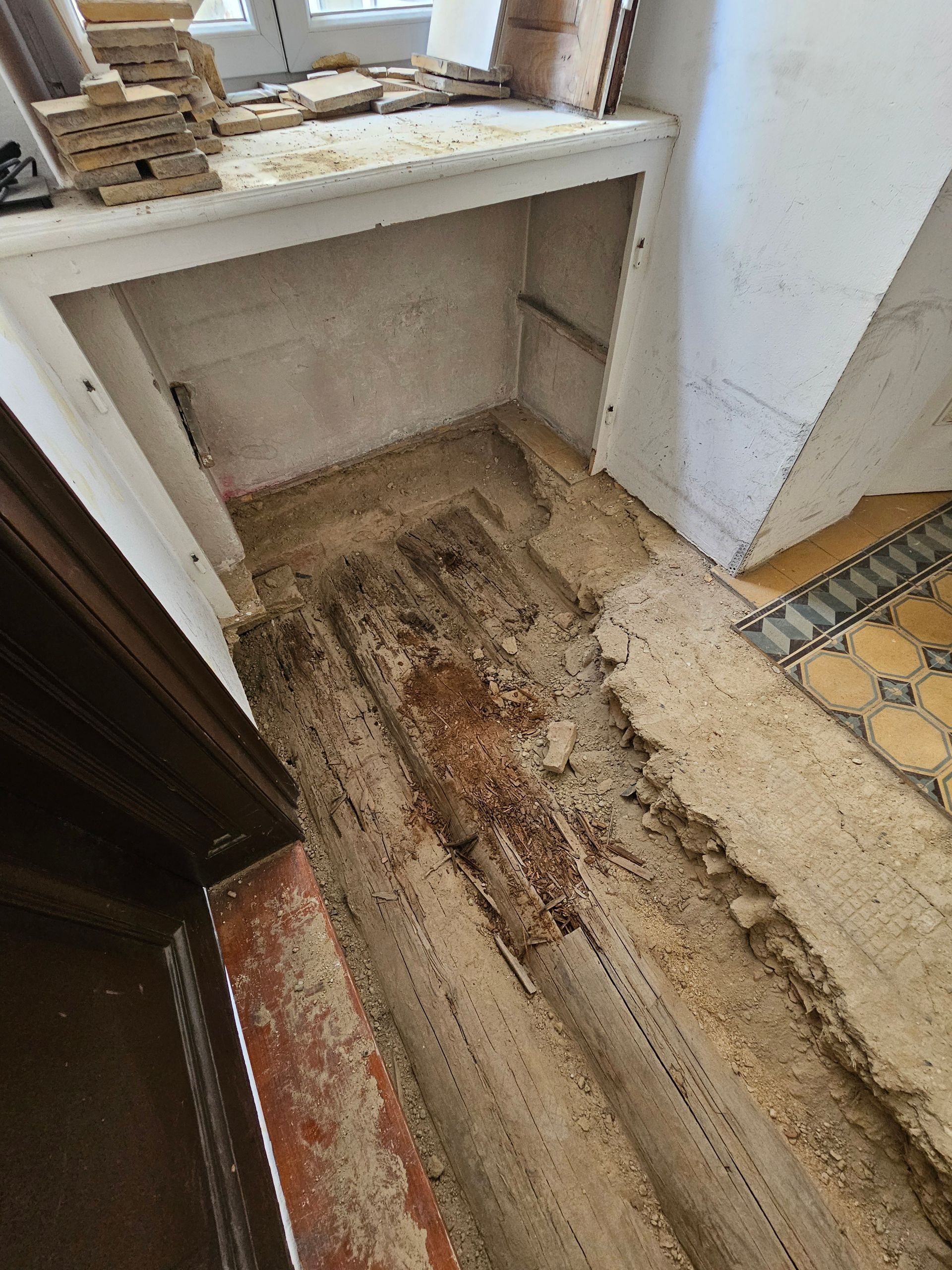 Ein beschädigter, unebener, mit Schmutz und Bauschutt bedeckter Boden auf einer Baustelle neben einem gefliesten Eingangsbereich und einem Holzregal.