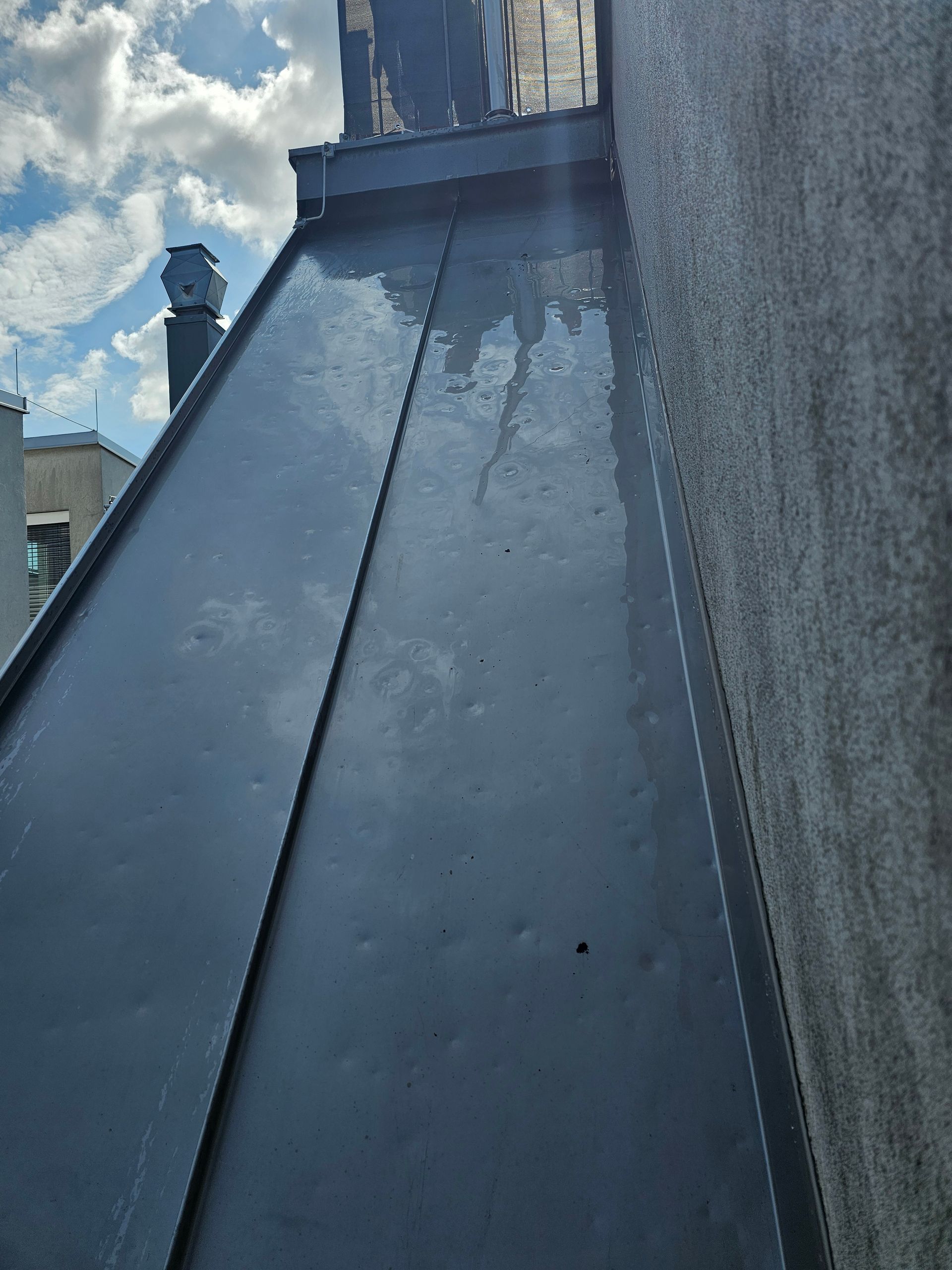 Blick hinunter auf einen schmalen, dunklen Metalldachabschnitt neben einer rauen, grauen Außenwand unter einem blauen, bewölkten Himmel.