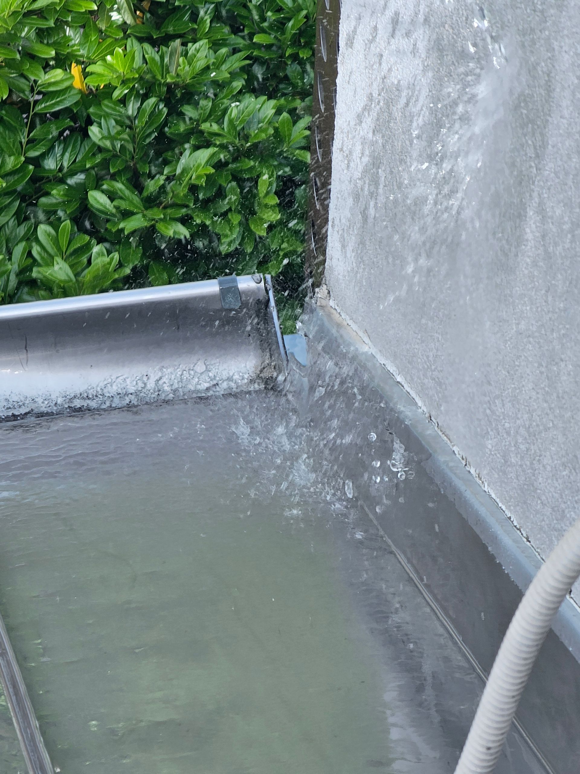 Bei starkem Regen spritzt Wasser von einer metallenen Dachrinnenverbindung in der Nähe einer grauen Außenwand; im Hintergrund ist grünes Laubwerk zu sehen.