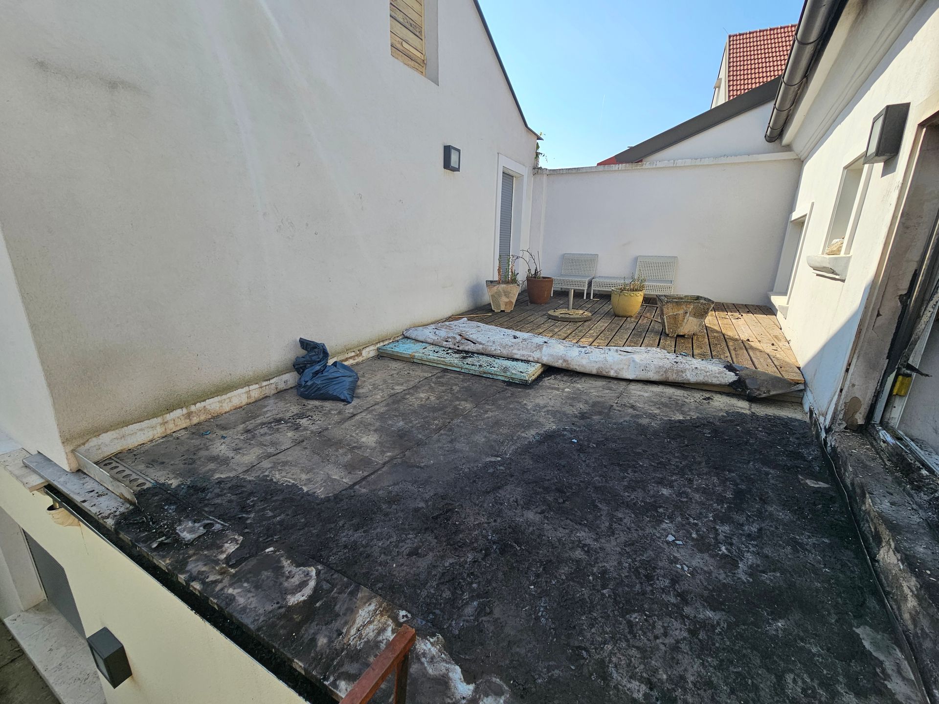 Eine Dachterrasse mit dunkler, fleckiger Oberfläche, die über eine kleine Sitzecke mit Töpfen und Kissen vor einer weißen Wand verfügt.