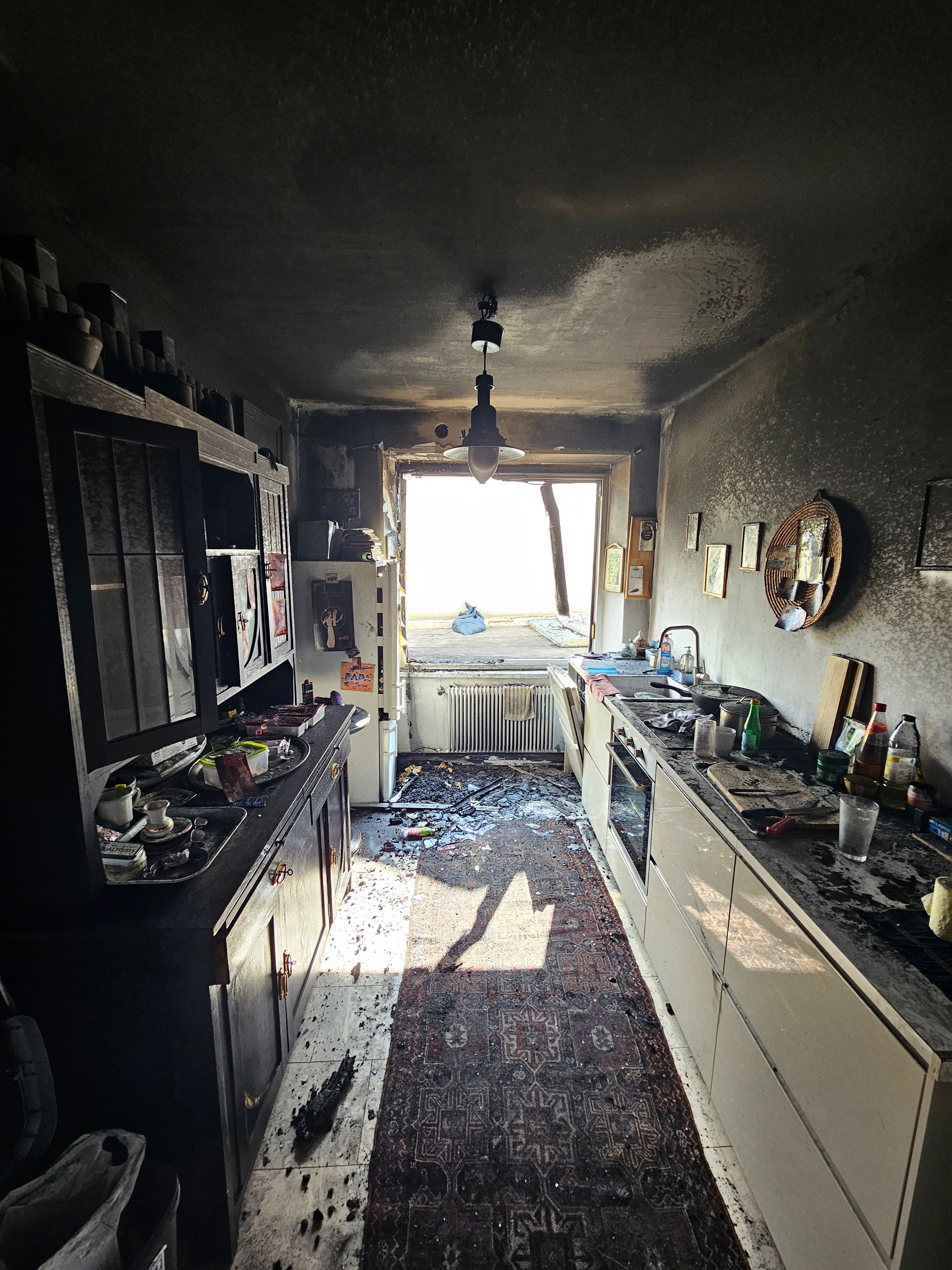 Eine schmale, rußbedeckte Küche nach einem Brand, auf der Trümmer auf dem Boden liegen und am anderen Ende eine helle, offene Tür zu sehen ist.