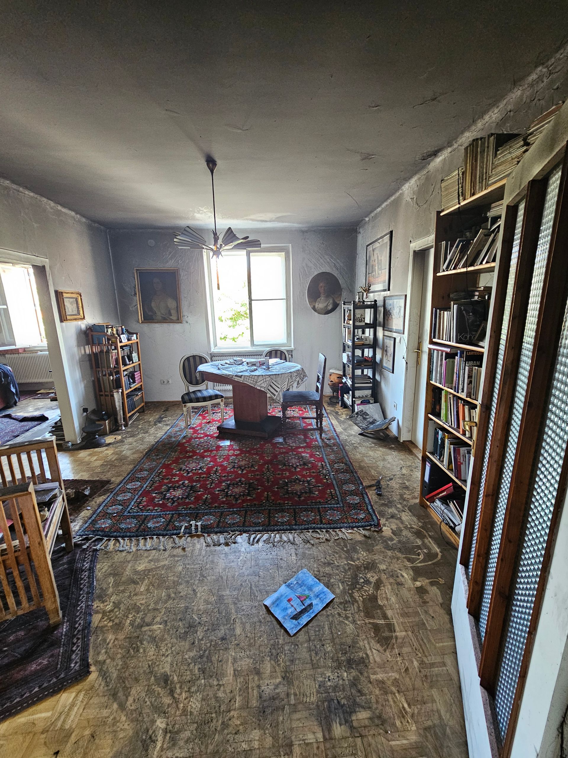 Ein Essbereich mit einem runden Tisch, einem rot gemusterten Teppich und hohen Bücherregalen, die sich unter einer schwach beleuchteten Deckenleuchte an der Wand entlangziehen.