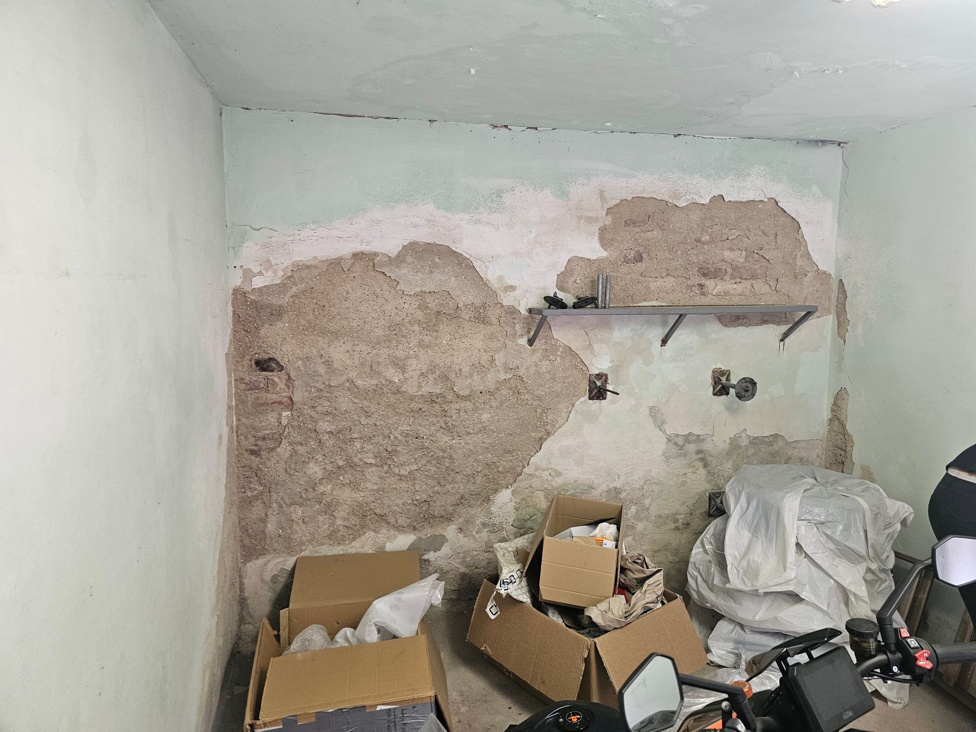 Eine Zimmerecke mit hellen Wänden, an denen der Putz abgebröckelt ist und das darunterliegende Mauerwerk freilegt, mit Kisten auf dem Boden.