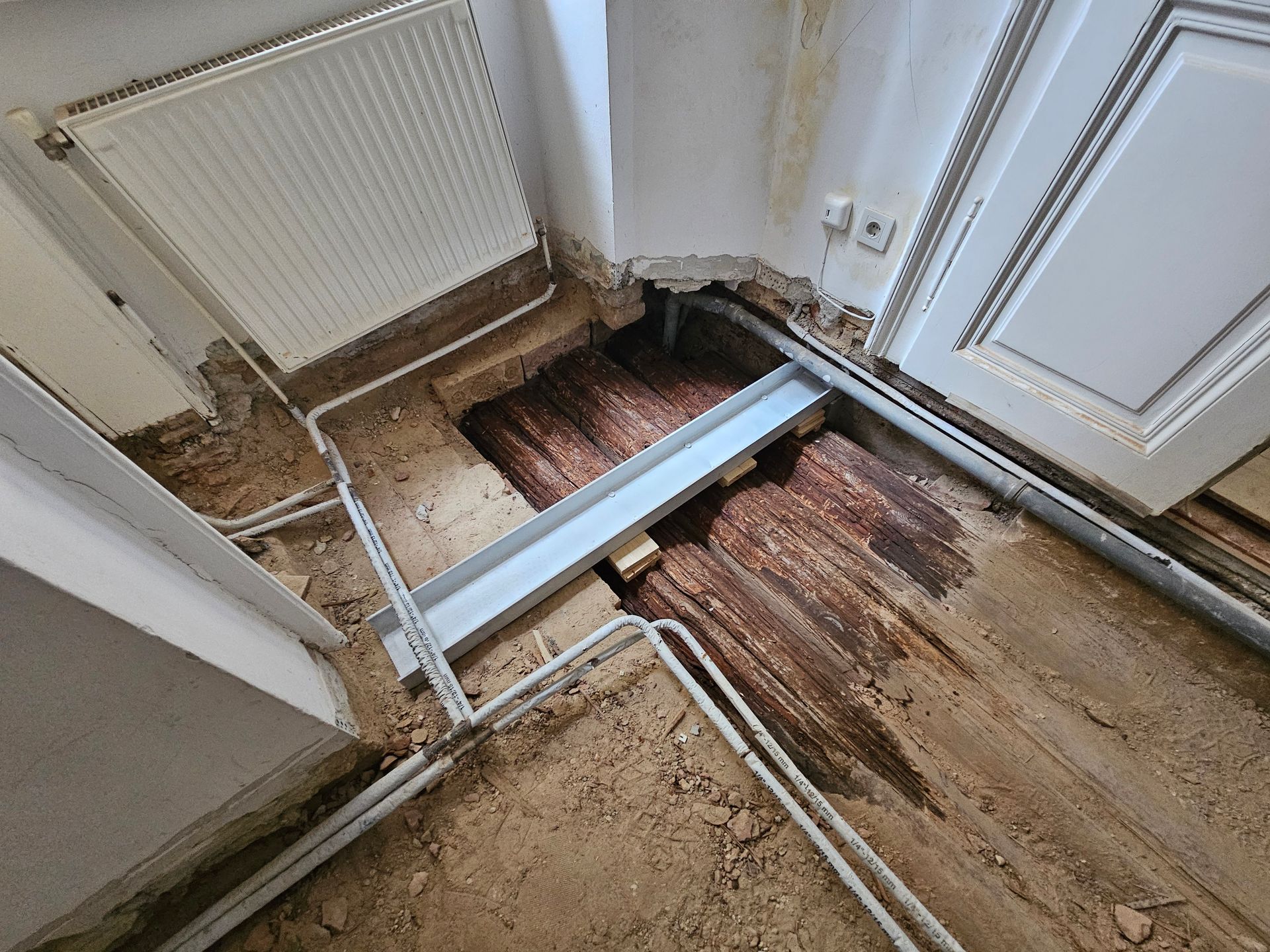 Ein Zimmer im Umbau mit freiliegenden Dielen, einer Metallbodenschiene, weißen Rohren und einem Heizkörper an der Wand.