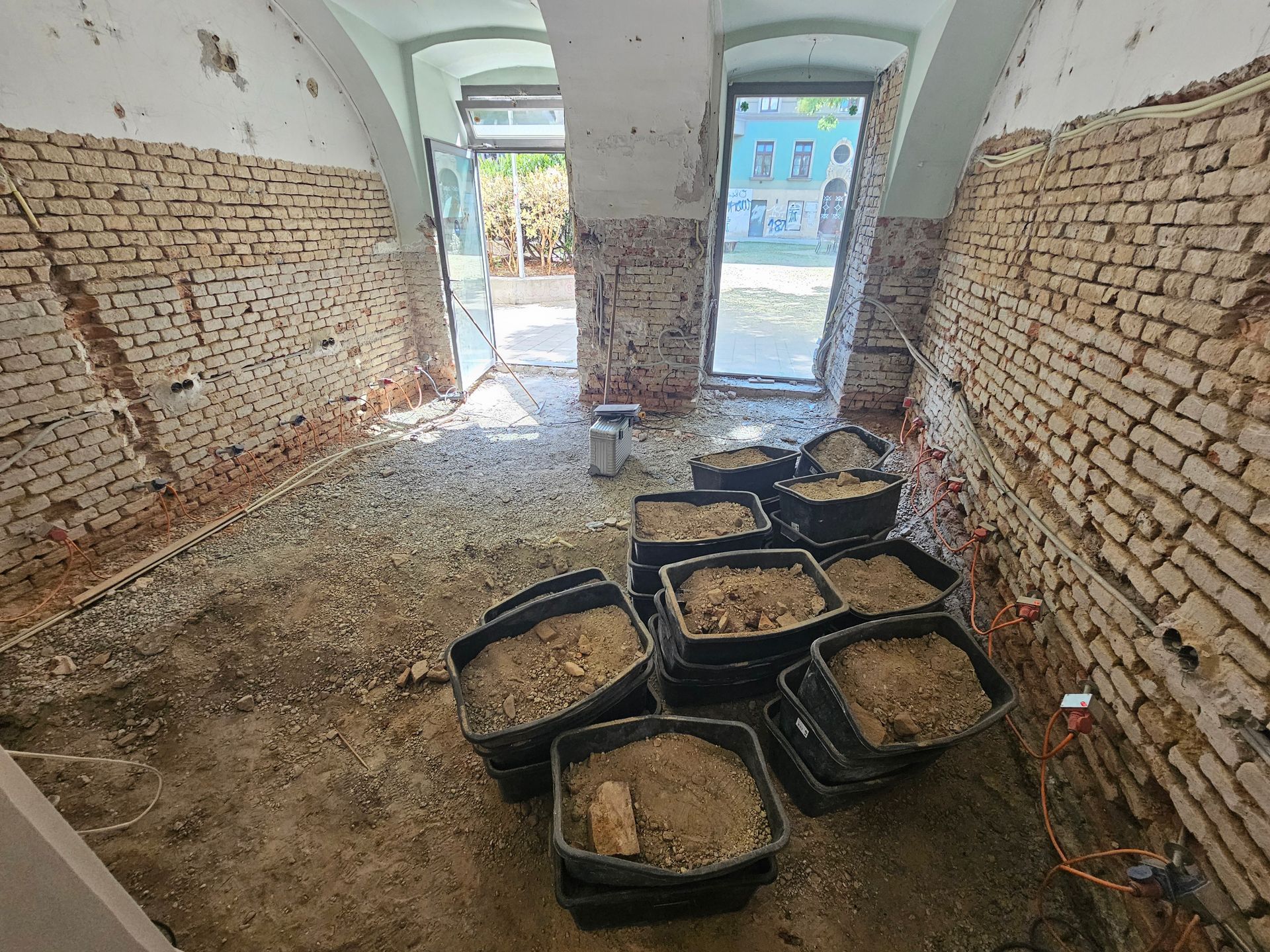 Ein unfertiger Kellerraum mit freiliegenden Ziegelwänden, einem Lehmboden und mehreren mit Bauschutt gefüllten Plastikbehältern.