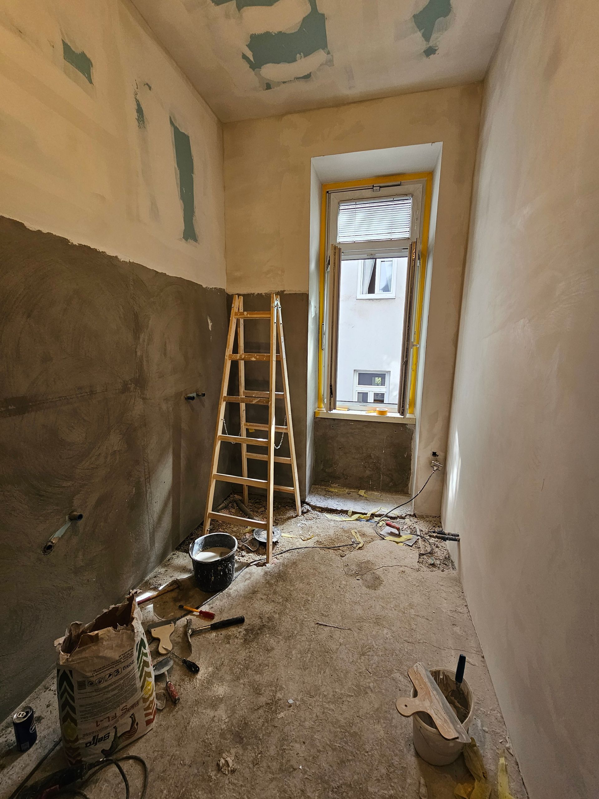 Ein Zimmer, das gerade renoviert wird, mit verputzten Wänden, einer Holzleiter, Bauschutt auf dem Boden und einem offenen Fenster.