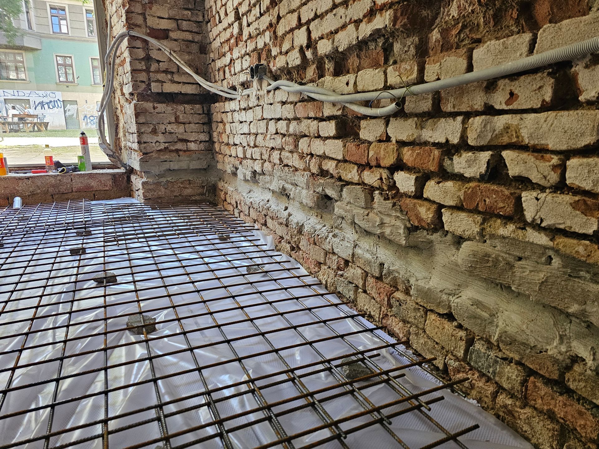 Eine Baustelle zeigt ein Drahtgitter über einer Kunststofffolie als Bodenbelag, angrenzend an eine freiliegende Ziegelwand.