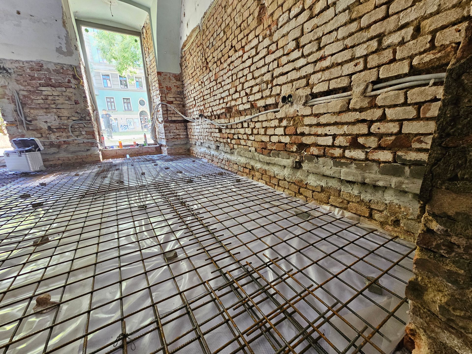 Im Bau befindlicher Raum mit Fußbodenheizungsrohren, die über einem Gitter auf weißem Blech an unverputzten Ziegelwänden verlegt sind.